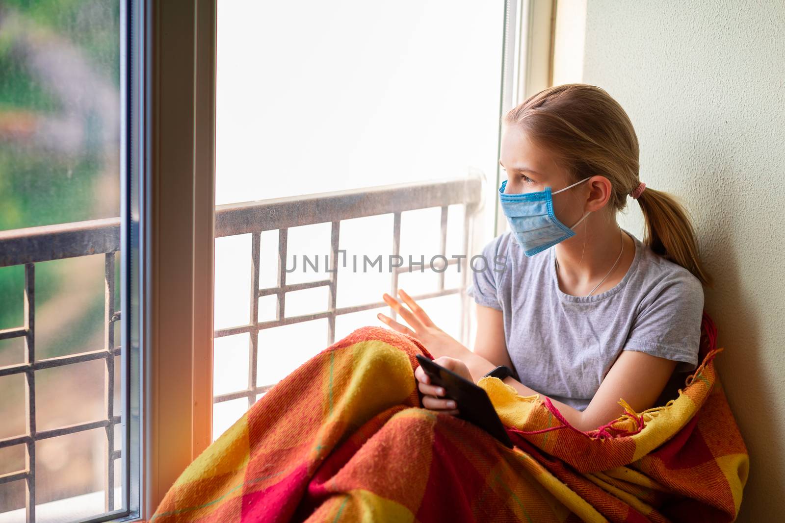 Sad little girl in protective mask sitting on the window with e-book or tablet reading and studying at home isolated. Coronavirus quarantine distance education concept.