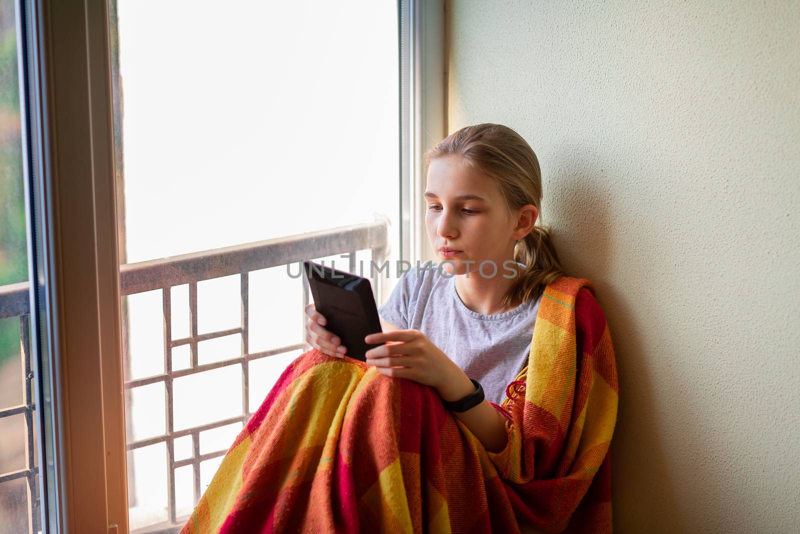 Sad little girl sitting on the window with e-book or tablet reading and studying at home isolated. Coronavirus quarantine distance education concept.