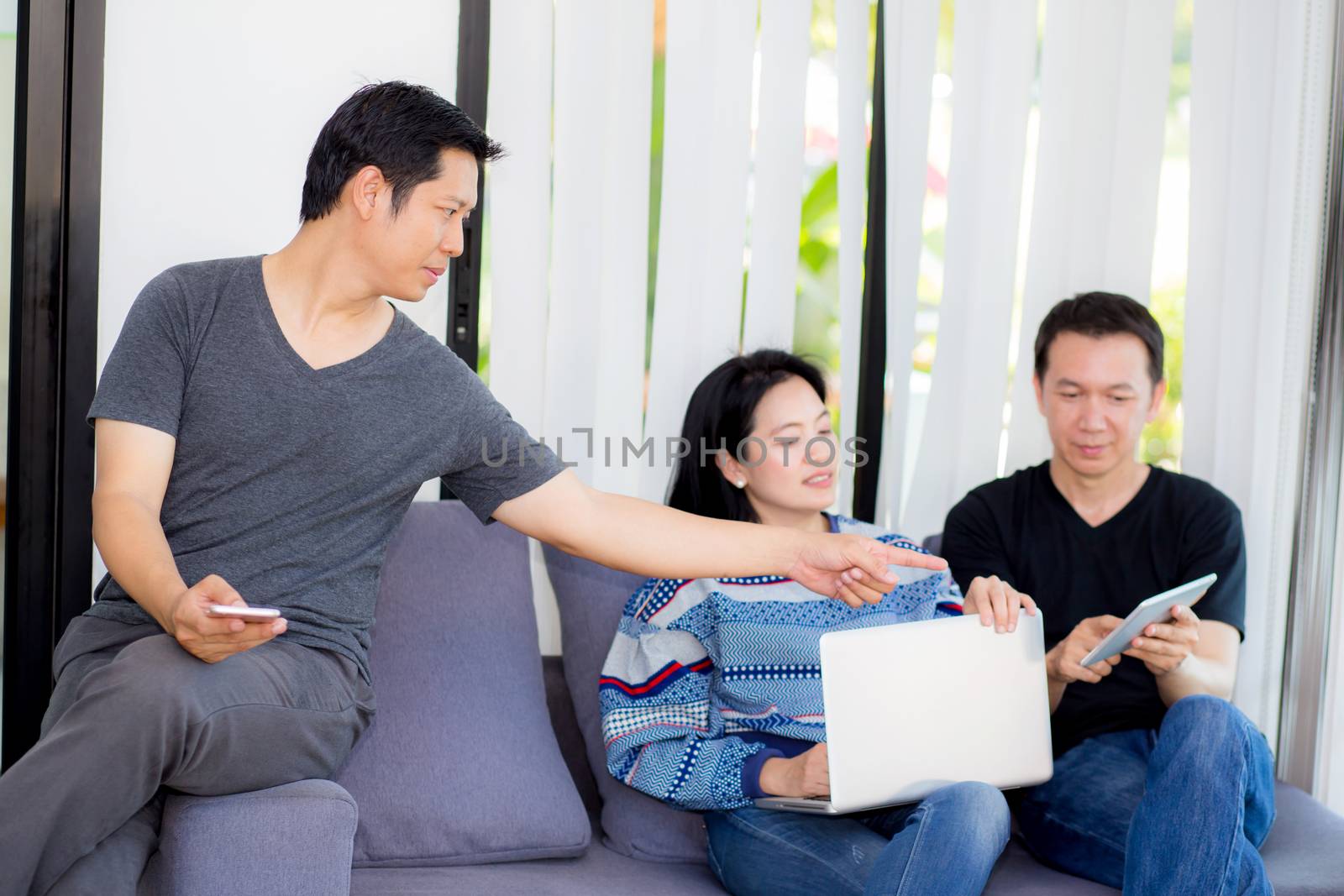 Three friends on line with multiple devices and talking sitting on a sofa in the living room in a house interior.