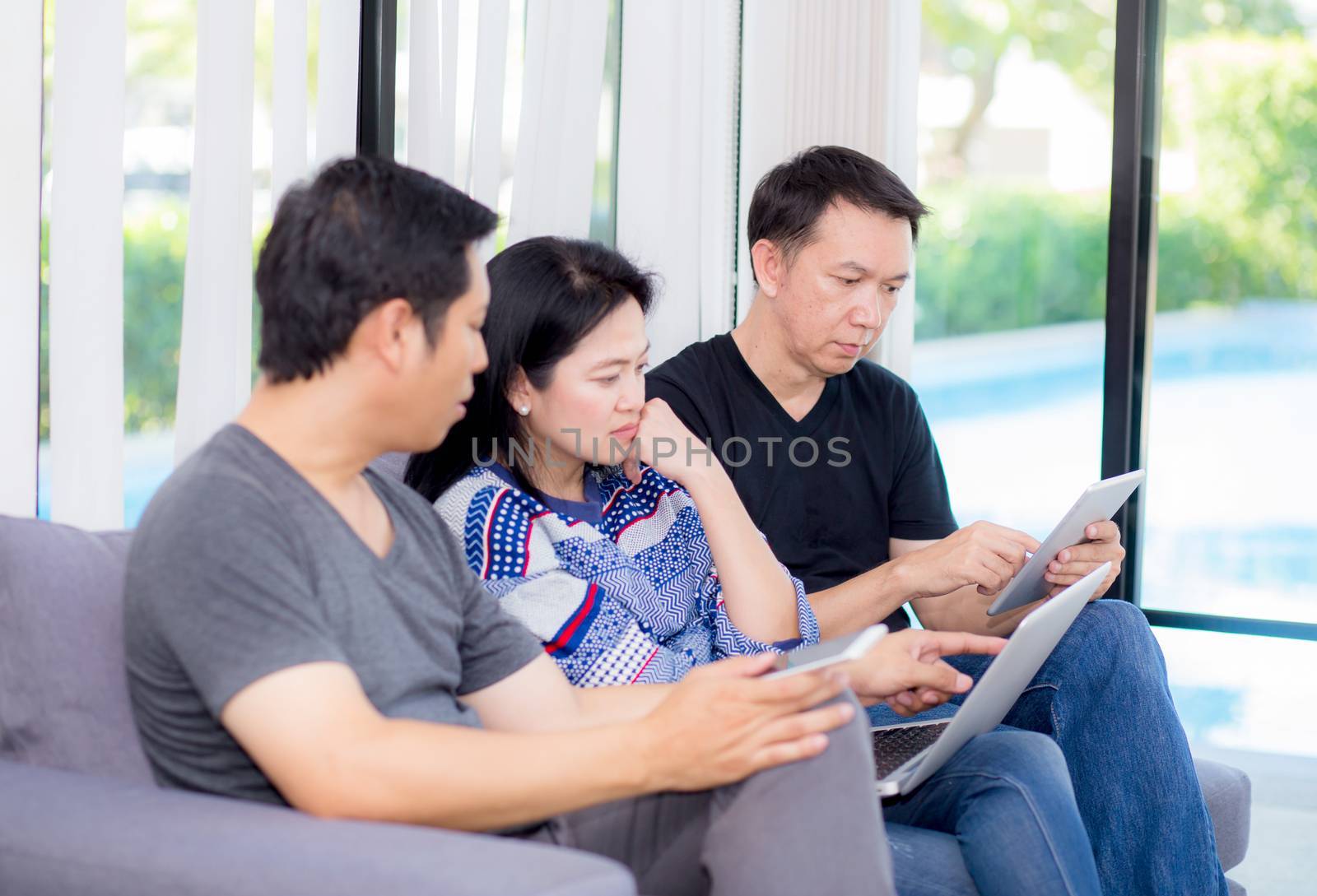 Three friends on line with multiple devices and talking sitting on a sofa in the living room in a house interior.