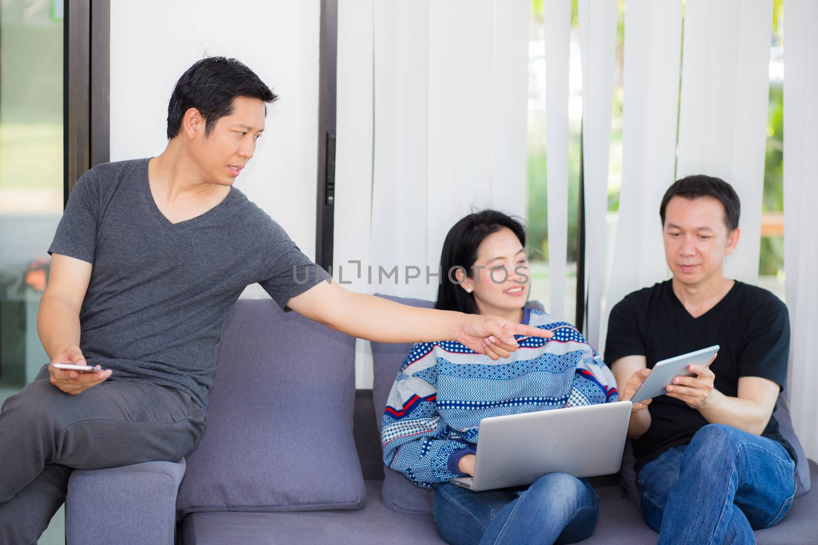 Three friends on line with multiple devices and talking sitting on a sofa in the living room in a house interior.