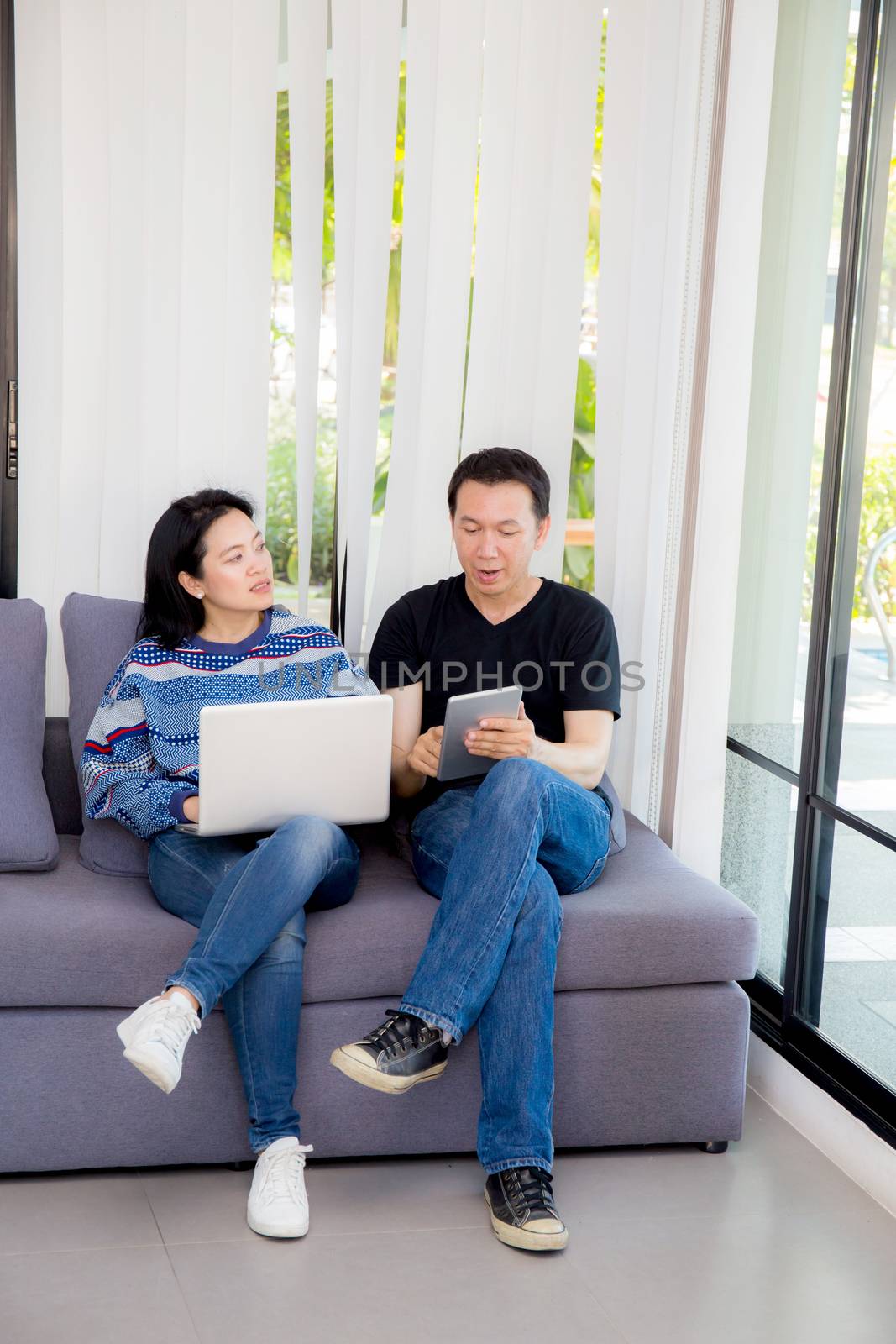 Two friends on line with multiple devices and talking sitting on a sofa in the living room in a house interior.
