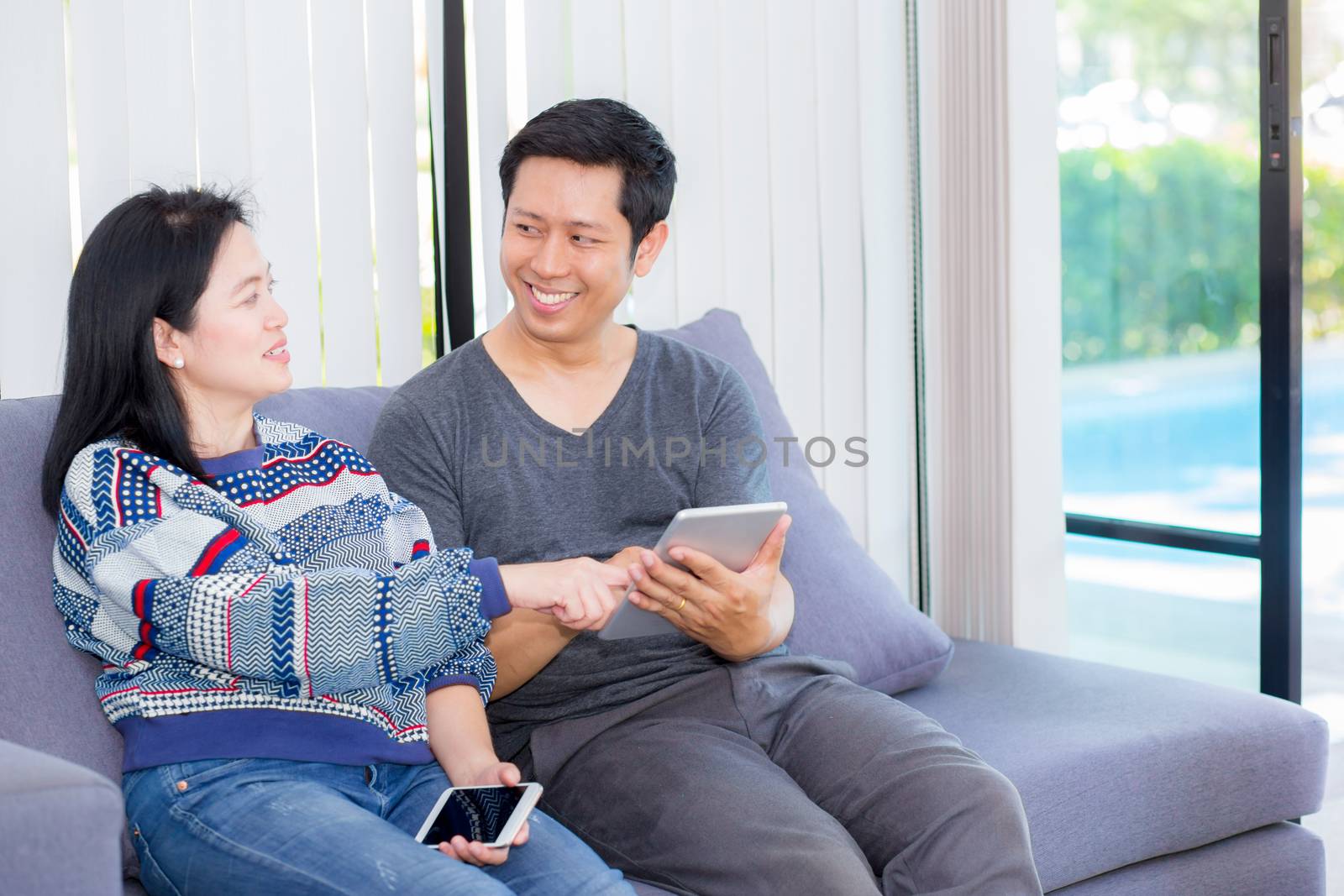 Two friends on line with multiple devices and talking sitting on a sofa in the living room in a house interior, communication concept.