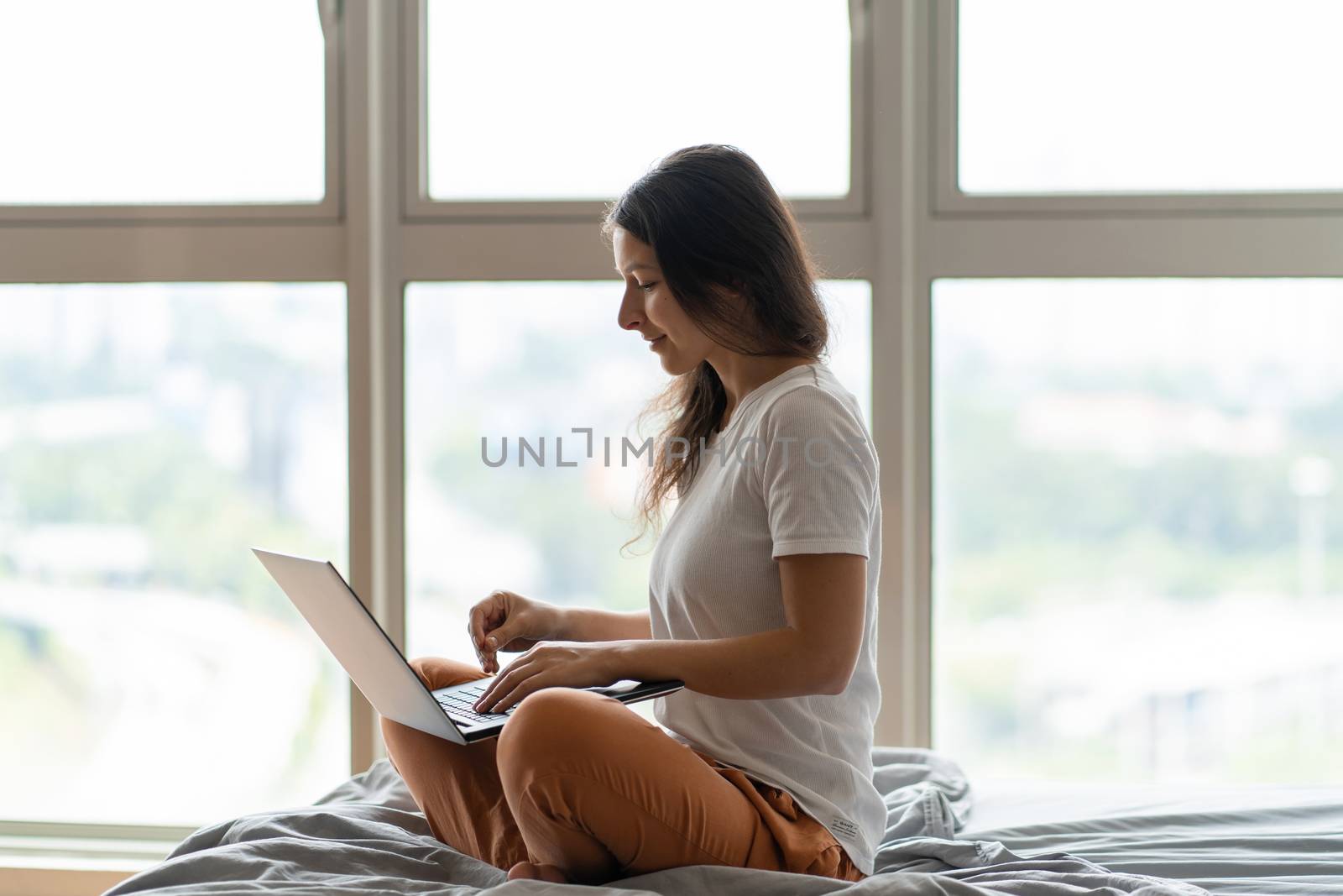 A beautiful young brunette girl is working on the laptop while sitting on a bed by a panoramic window with a beautiful view from a high floor. Stylish modern interior. Shopping on the Internet. by Try_my_best