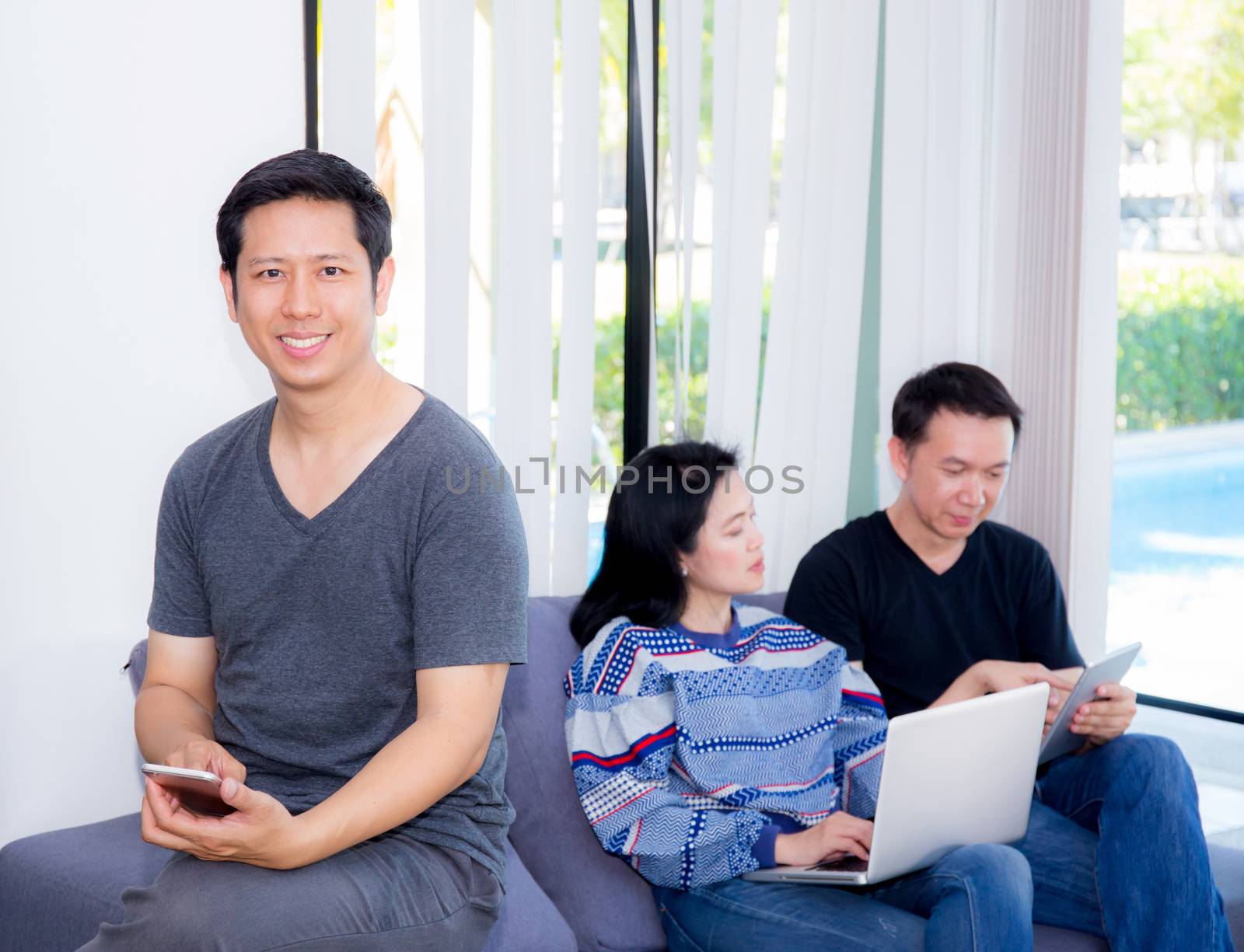 Three friends on line with multiple devices and talking sitting on a sofa in the living room in a house interior.