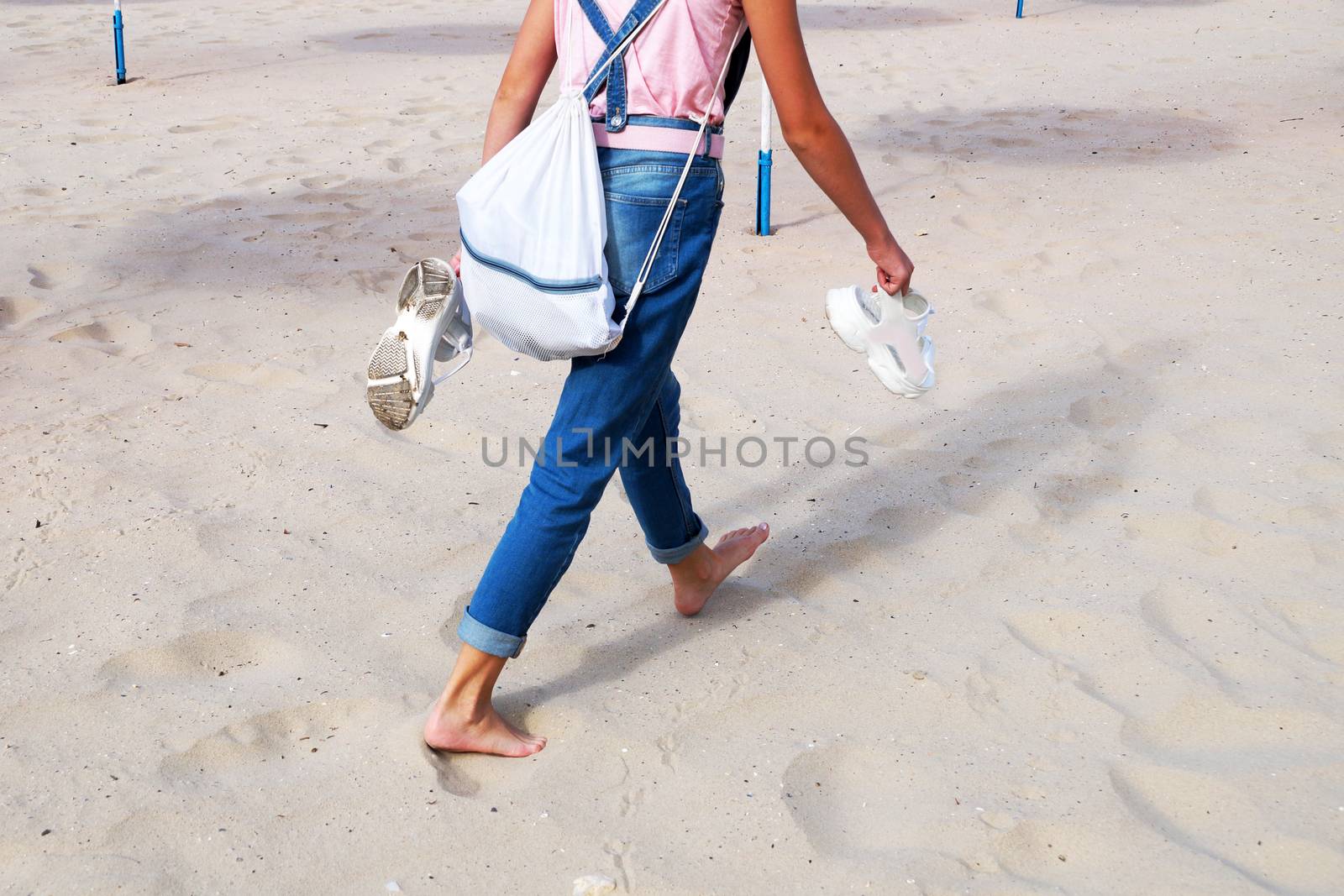a girl in jeans walks on the sand barefoot, holding sandals in her hands, rear view. by Annado