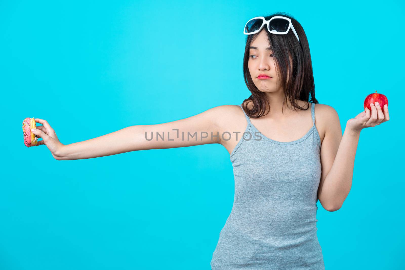 Attractive Asian young woman holding and choosing between disk of donuts or red apple fruit on isolated blue color background, weight loss and avoid junk food for dieting and Healthy