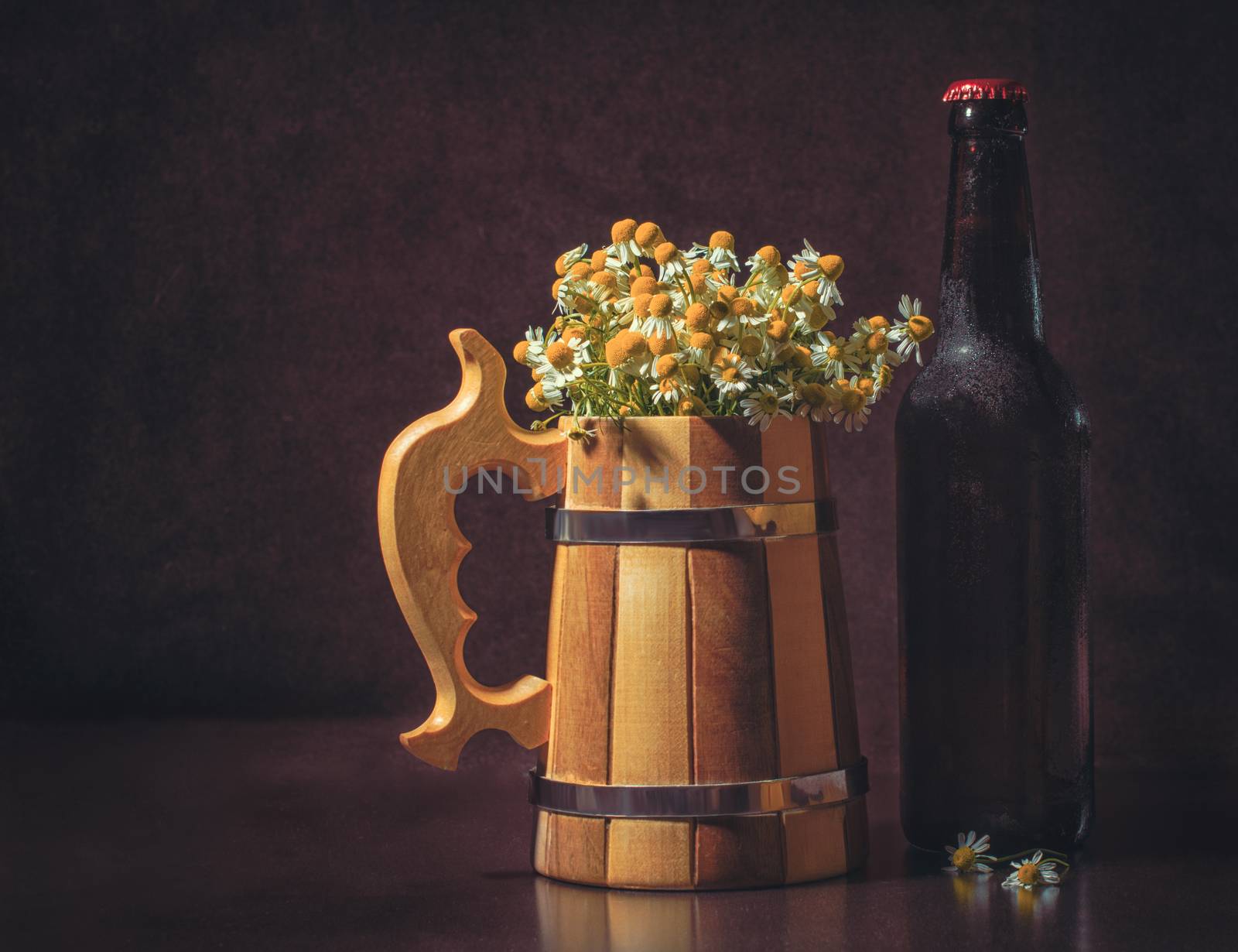 still life with a bottle of beer with a wooden mug and camomiles imitating foam on a dark brown background