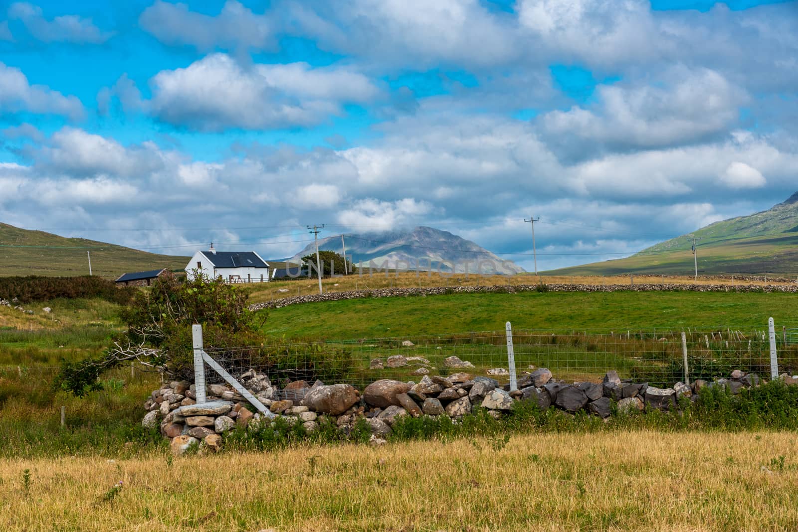 A farmhouse sits on a hill under a blue sky. A stone wall and fencing are in the foreground; a mountain is in the background.