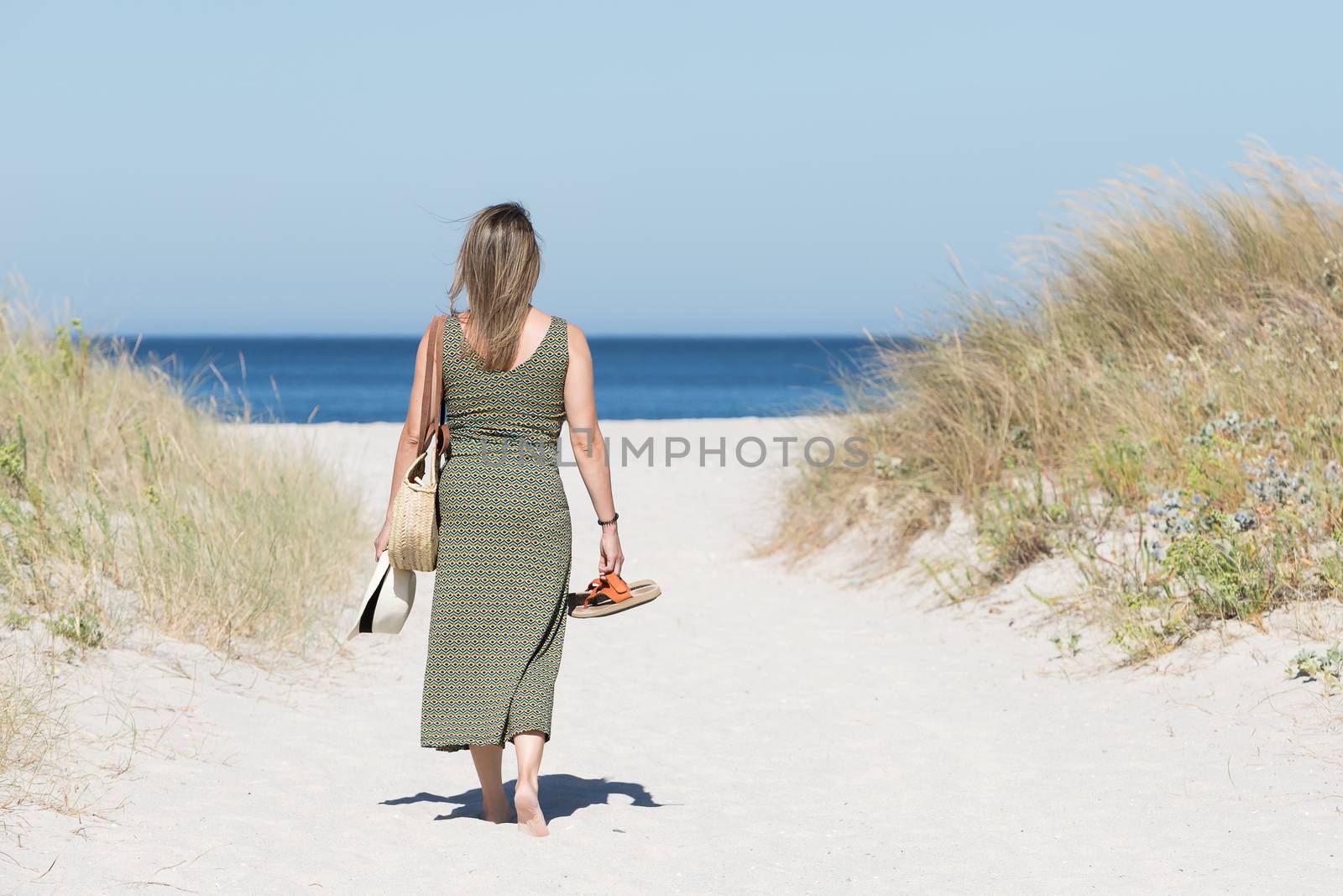 Middle-aged blonde woman in a dress walking barefoot along a sandy path towards the sea with her sandals in one hand and a hat in the other.
