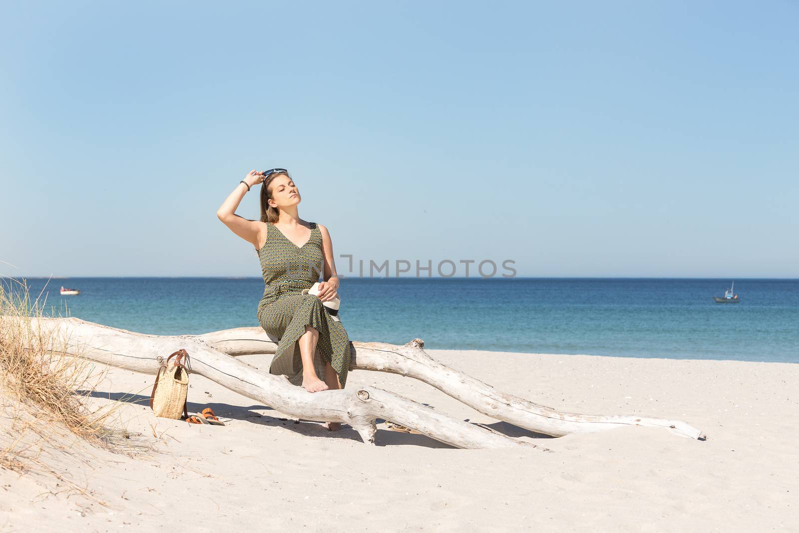 Middle-aged blonde woman sitting on a tree trunk wearing sunglasses on her head with the sea and the beach on her back