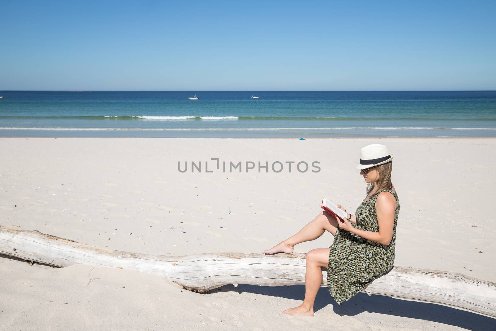 Middle-aged blonde woman sitting on a tree trunk n the beach reading a book with the sea on the background
