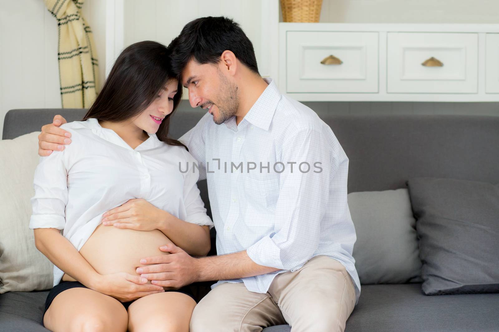 Beautiful pregnant asian woman and husband together care her pregnant on a sofa, Expectant parents, Family concept.