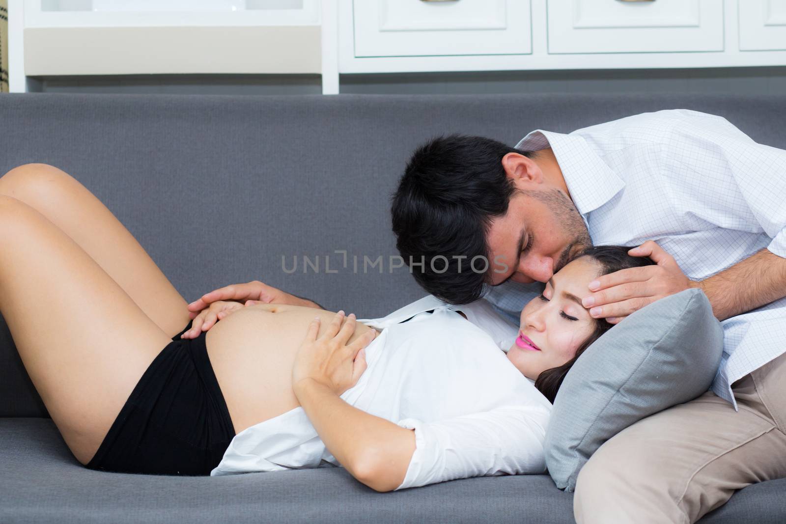 Husband kissing forehead of wife with pregnant belly of while sitting on sofa.