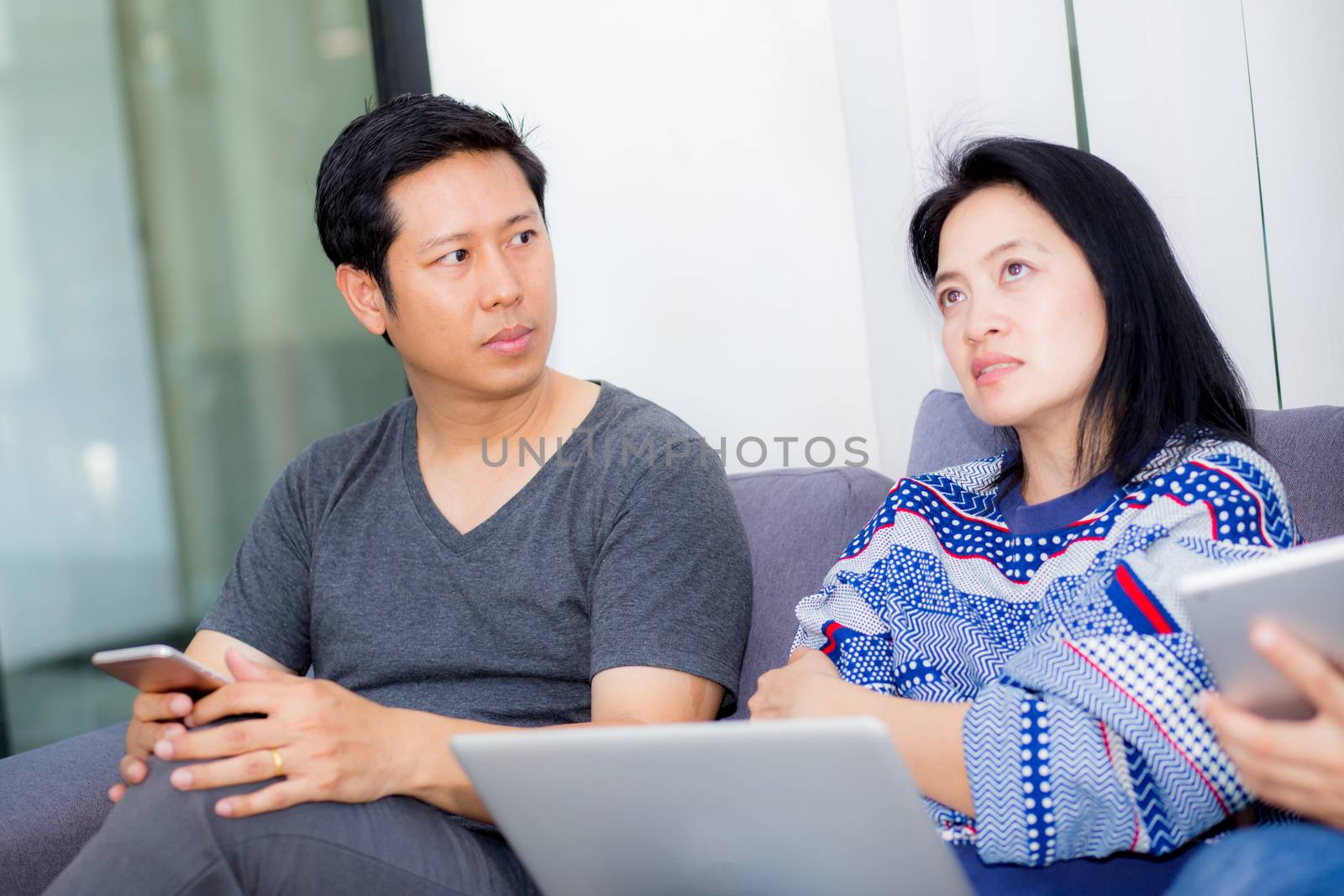 Asian two people friends on line with multiple devices and talking sitting on a sofa in the living room in a house interior, communication concept.