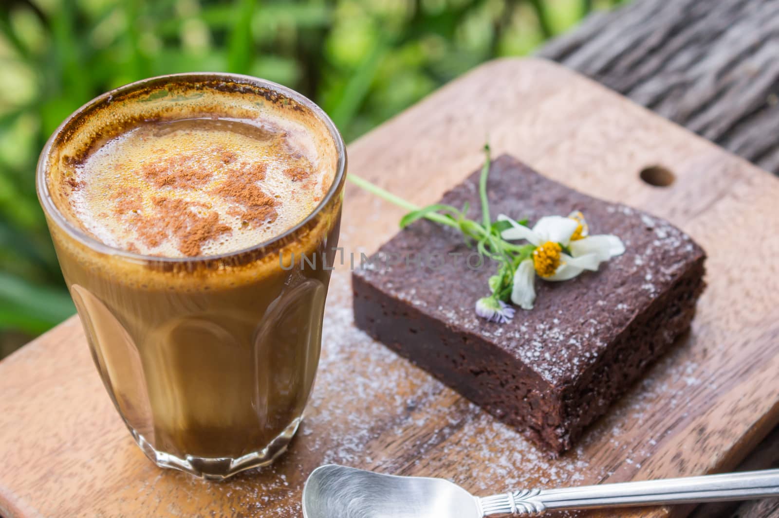 Chocolate Brownie Cake and Latte Coffee and Daisy Flower on Chopping Board or Cutting Board on Wood Table on Green Tree Background Top Frame.