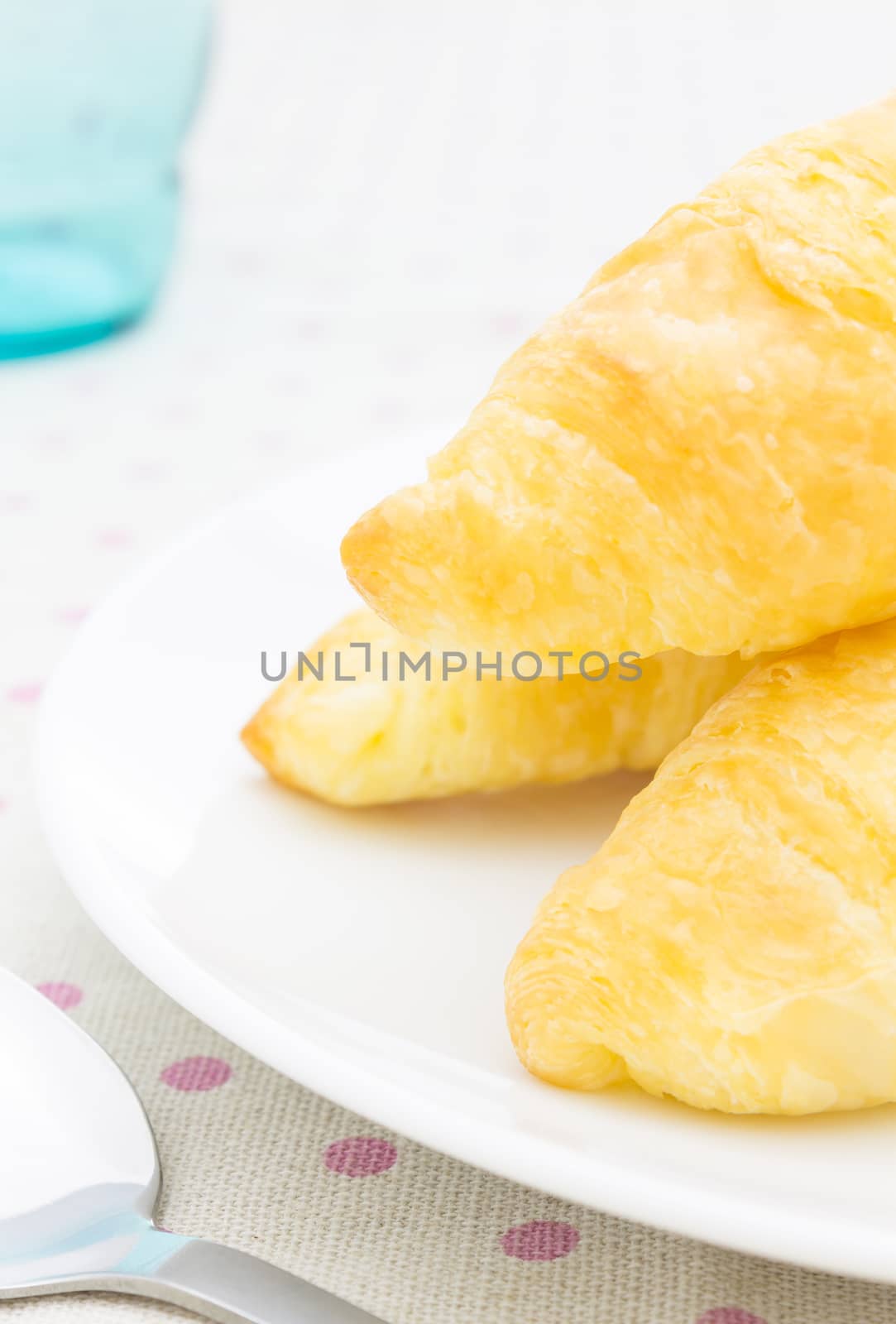 Croissant or bread or bakeray on white dish on pink point placemat with spoon and glass close up view