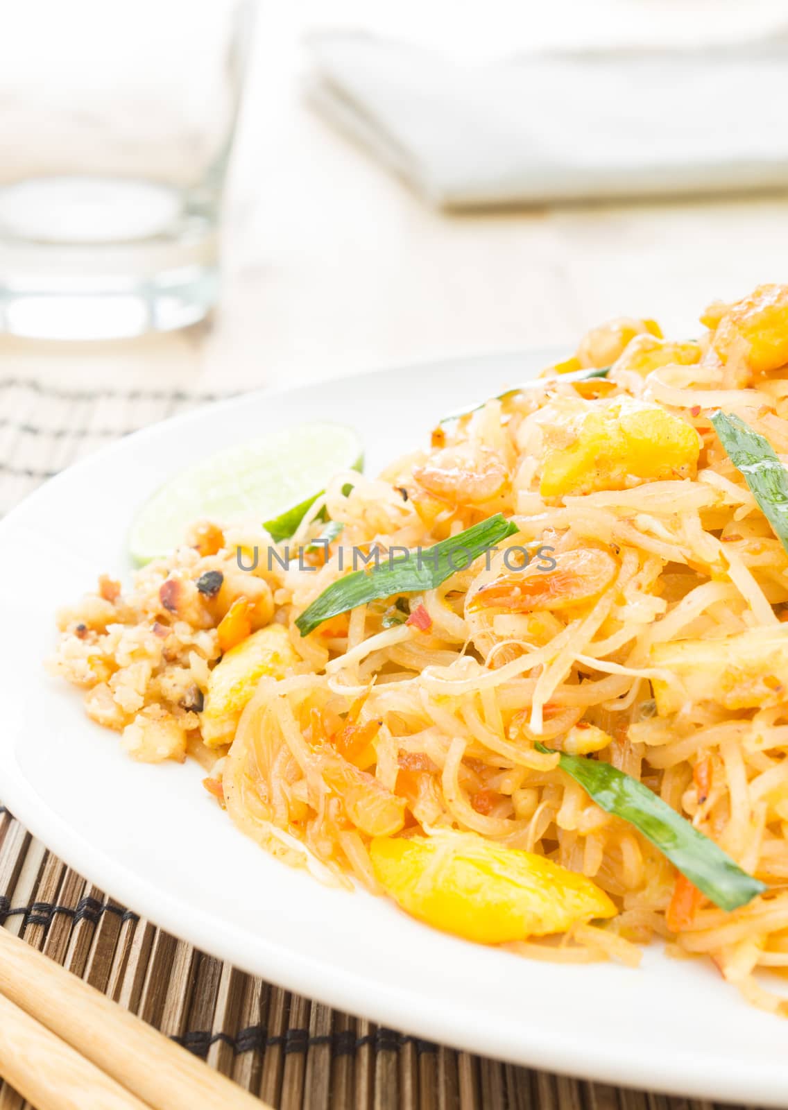 Pad Thai or Thai fried noodles on white dish on bamboo placemats vertical style. Glass and napkin chopsticks egg dry shrimps onions sprouts lemon peanut