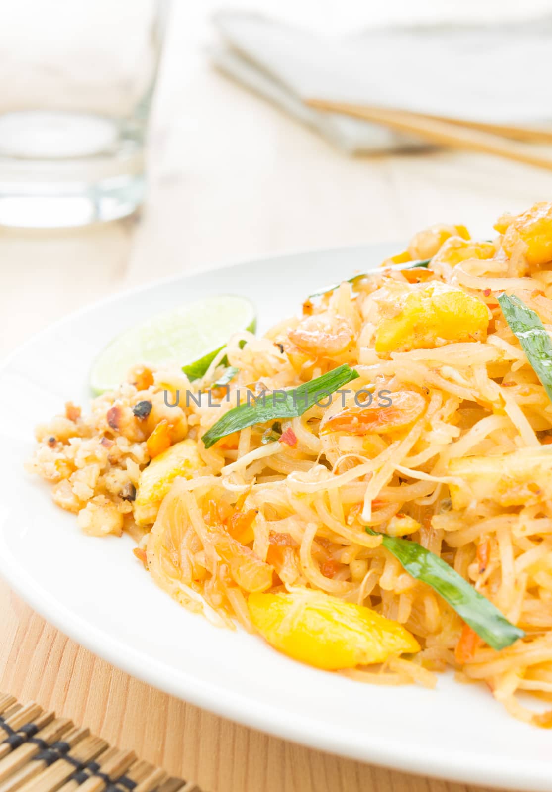 Pad Thai or Thai fried noodles on white dish on wood table vertical style. Glass and napkin chopsticks egg dry shrimps onions sprouts lemon peanut