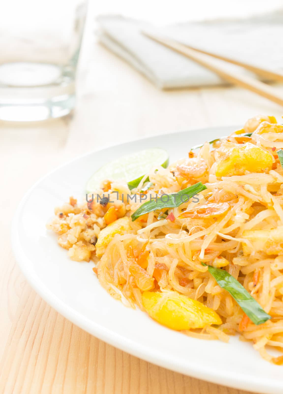 Pad Thai or Thai fried noodles on white dish on wood table vertical style. Glass and napkin chopsticks egg dry shrimps onions sprouts lemon peanut