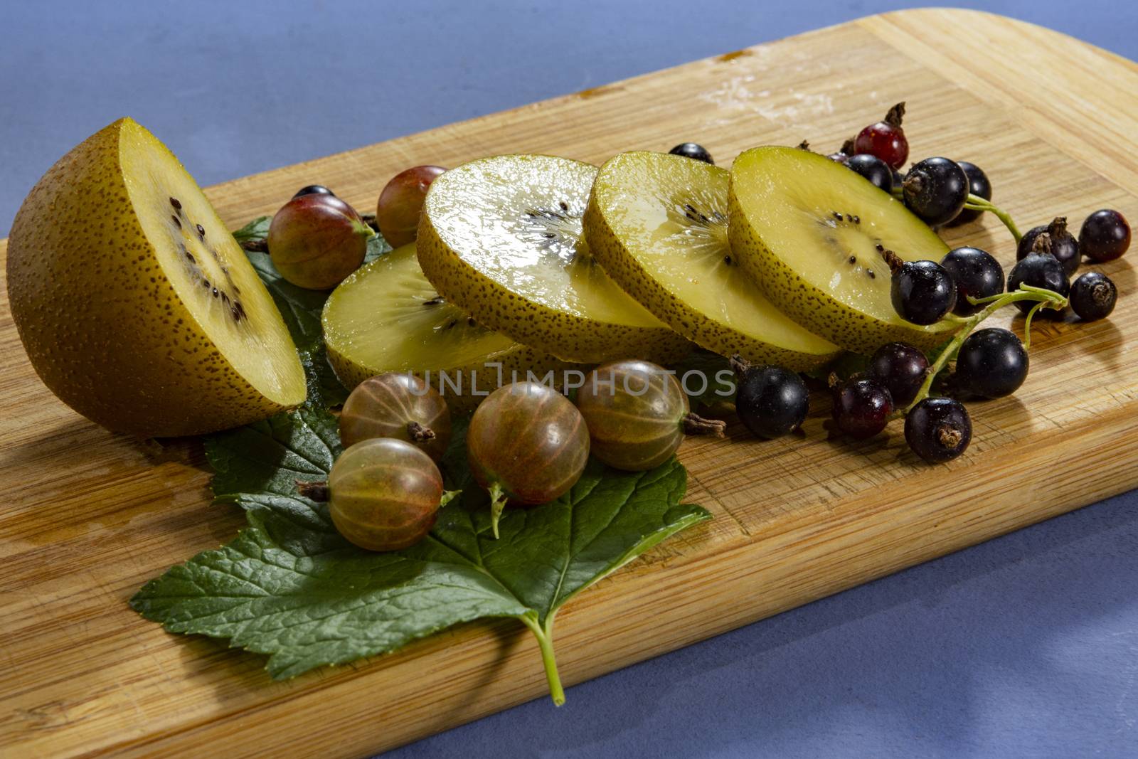 Kiwi sliced, gooseberries and black currants with leaves on a wooden board on a blue  background