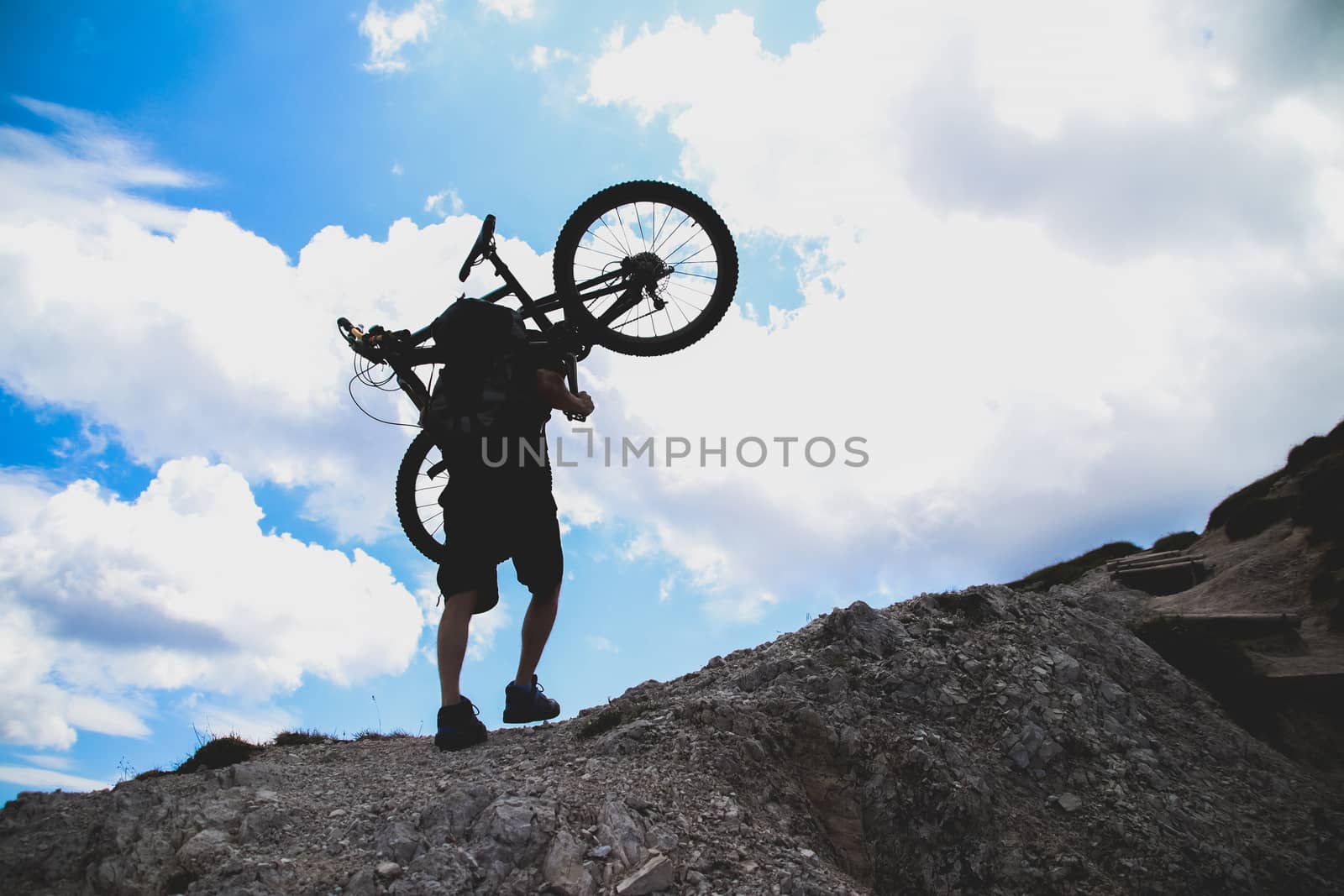 sport man in action lifting bike on his shoulder on rock mountai by anotestocker