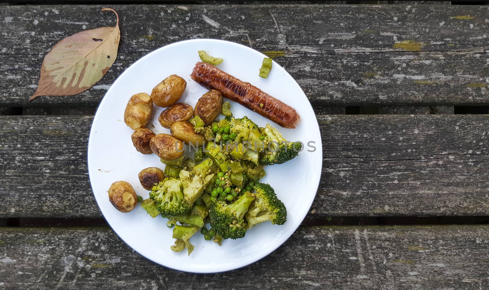 Vegan meal with oven baked potatos, plant based sausace, broccoli, peas and pesto on a white plate. Veganism and delicious food on weathered wooden table..