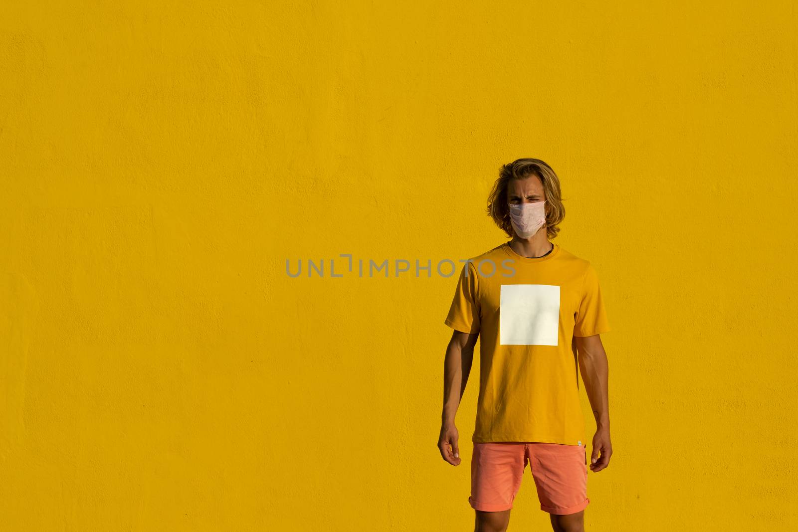 Blond young man with mask on a yellow background. Summer coronavirus concept