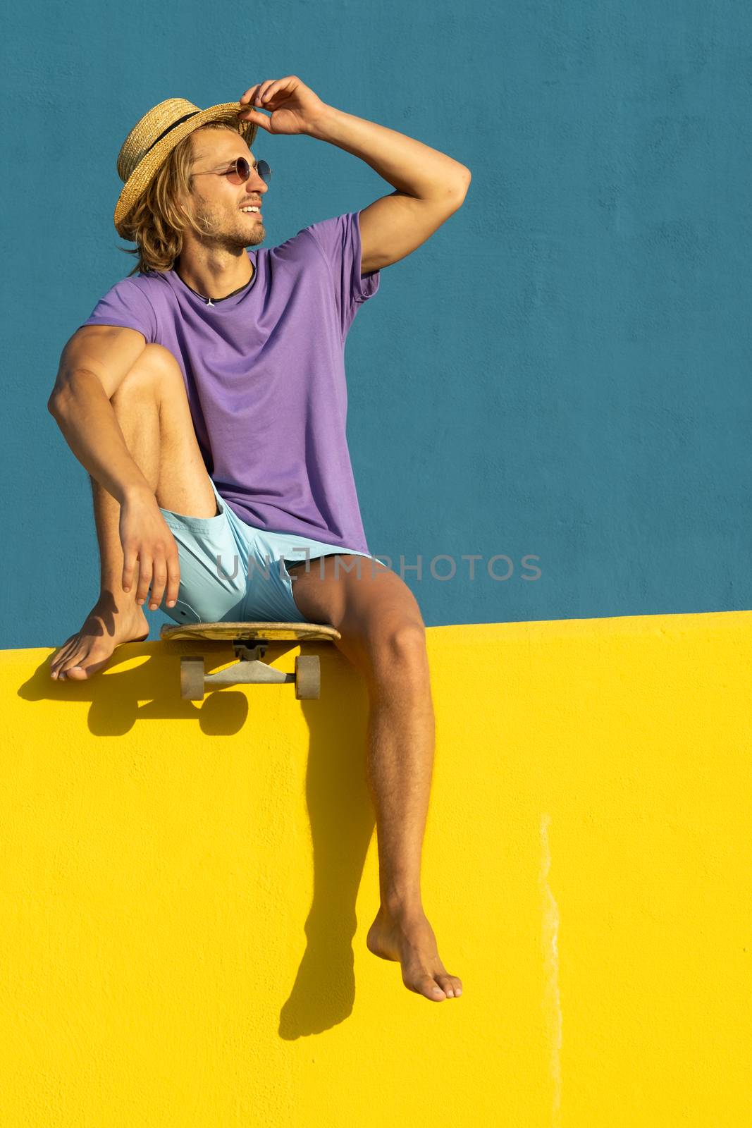 Blond young man with skateboard, hat and sunglasses enjoying the summer