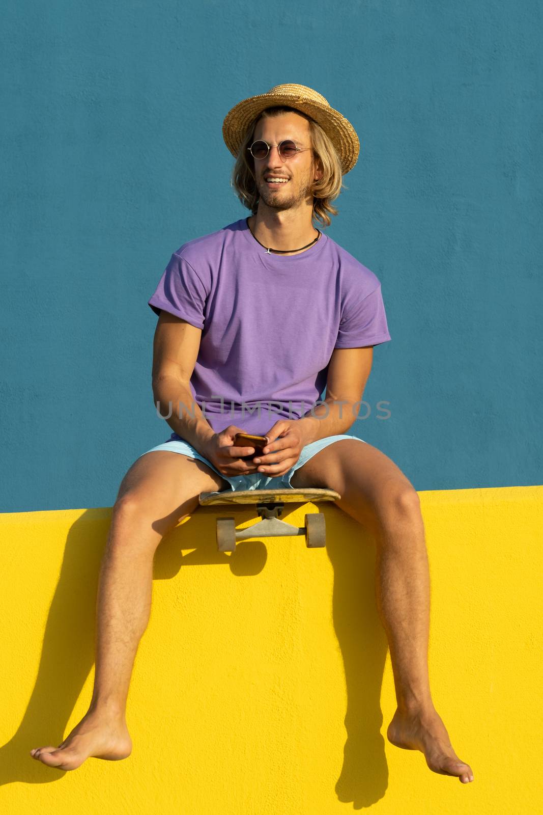 Blond young man with skateboard, mobile phone, hat and sunglasses enjoying the summer. Taking a selfie