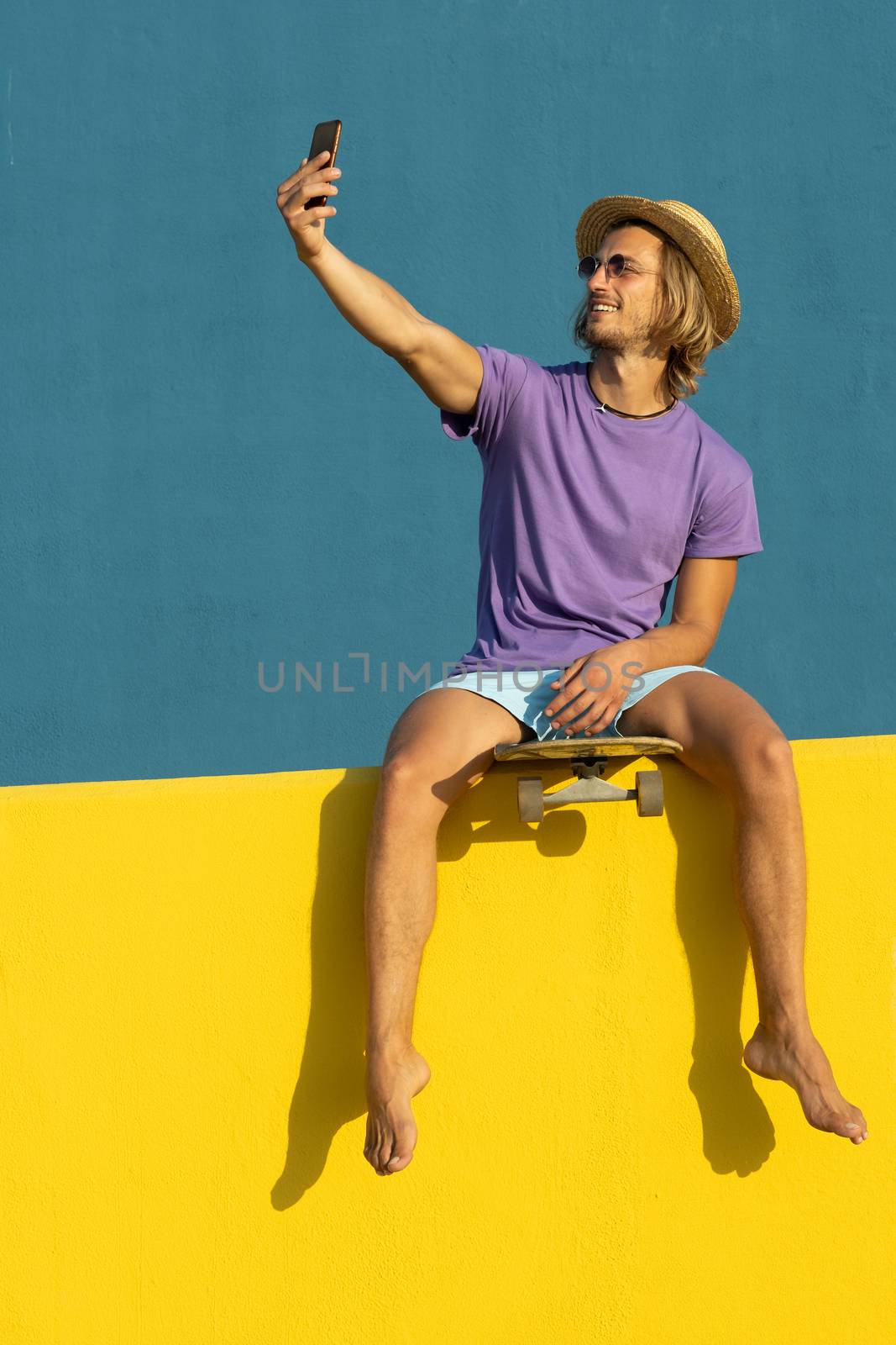 Blond young man with skateboard, mobile phone, hat and sunglasses enjoying the summer. Taking a selfie