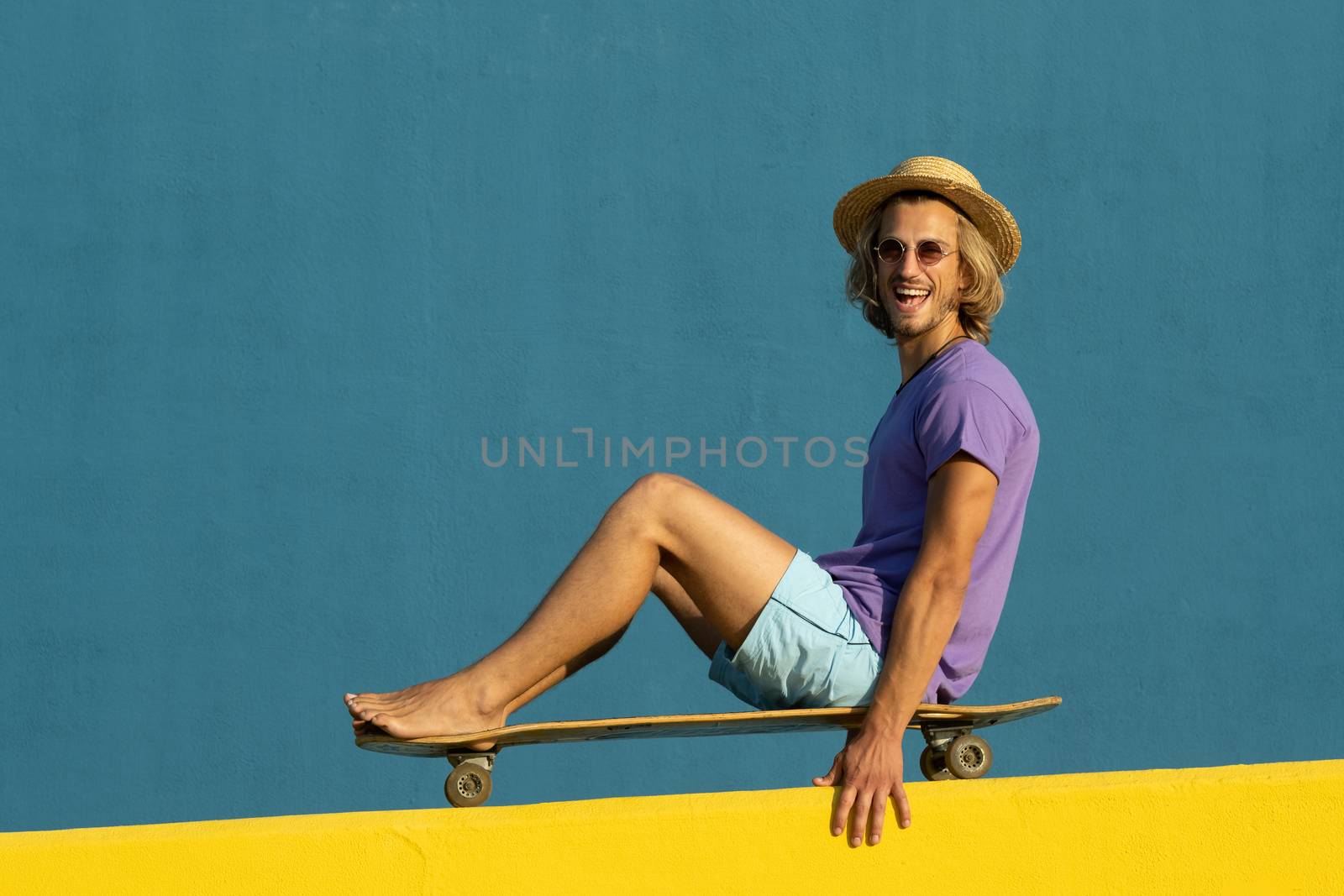 Blond young man with skateboard, hat and sunglasses enjoying the summer