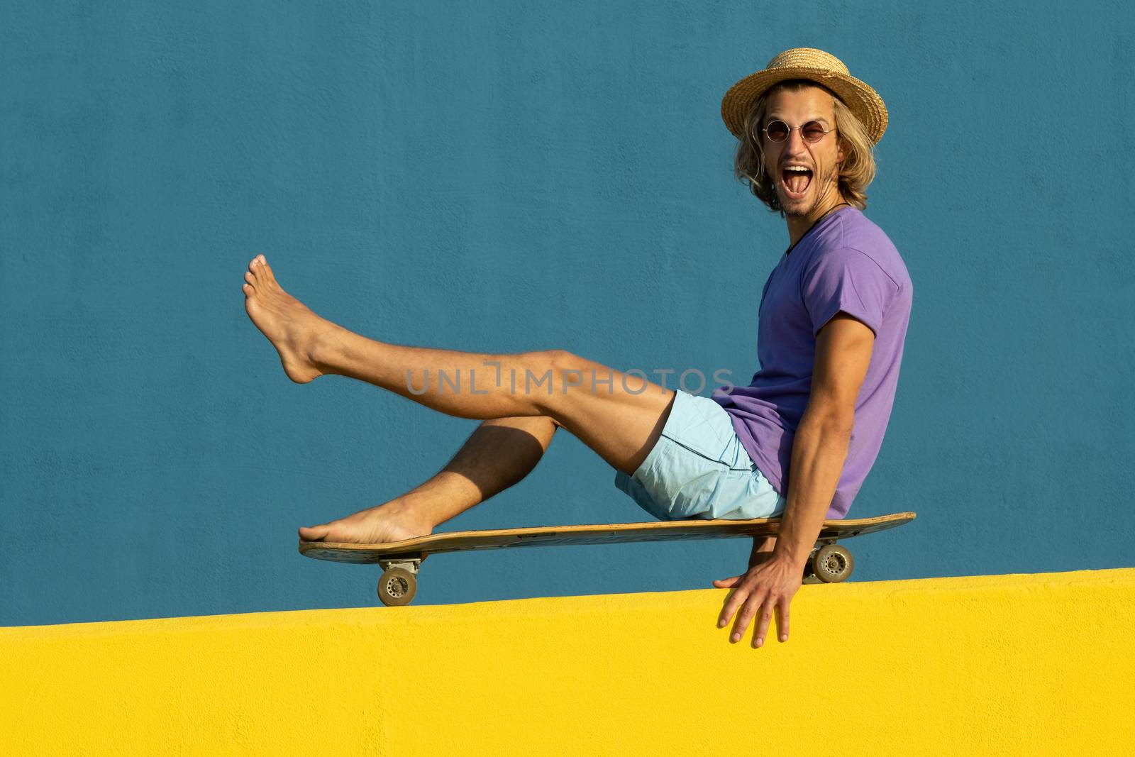 Blond young man with skateboard, hat and sunglasses enjoying the summer