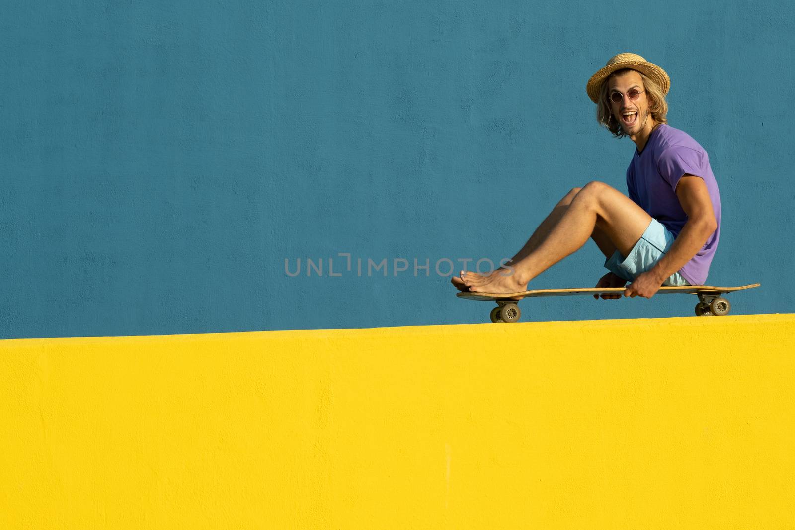 Blond young man with skateboard, hat and sunglasses enjoying the summer
