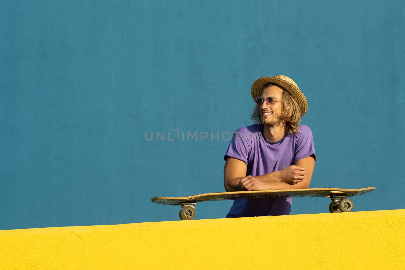 Blond young man with skateboard, hat and sunglasses enjoying the summer