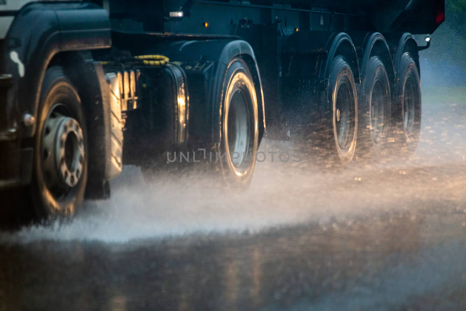 Rain water splash flow from wheels of heavy truck moving fast in daylight cit. Truck moving on asphalt road during heavy summer storm rain. Close-up with selective focus and blur.
