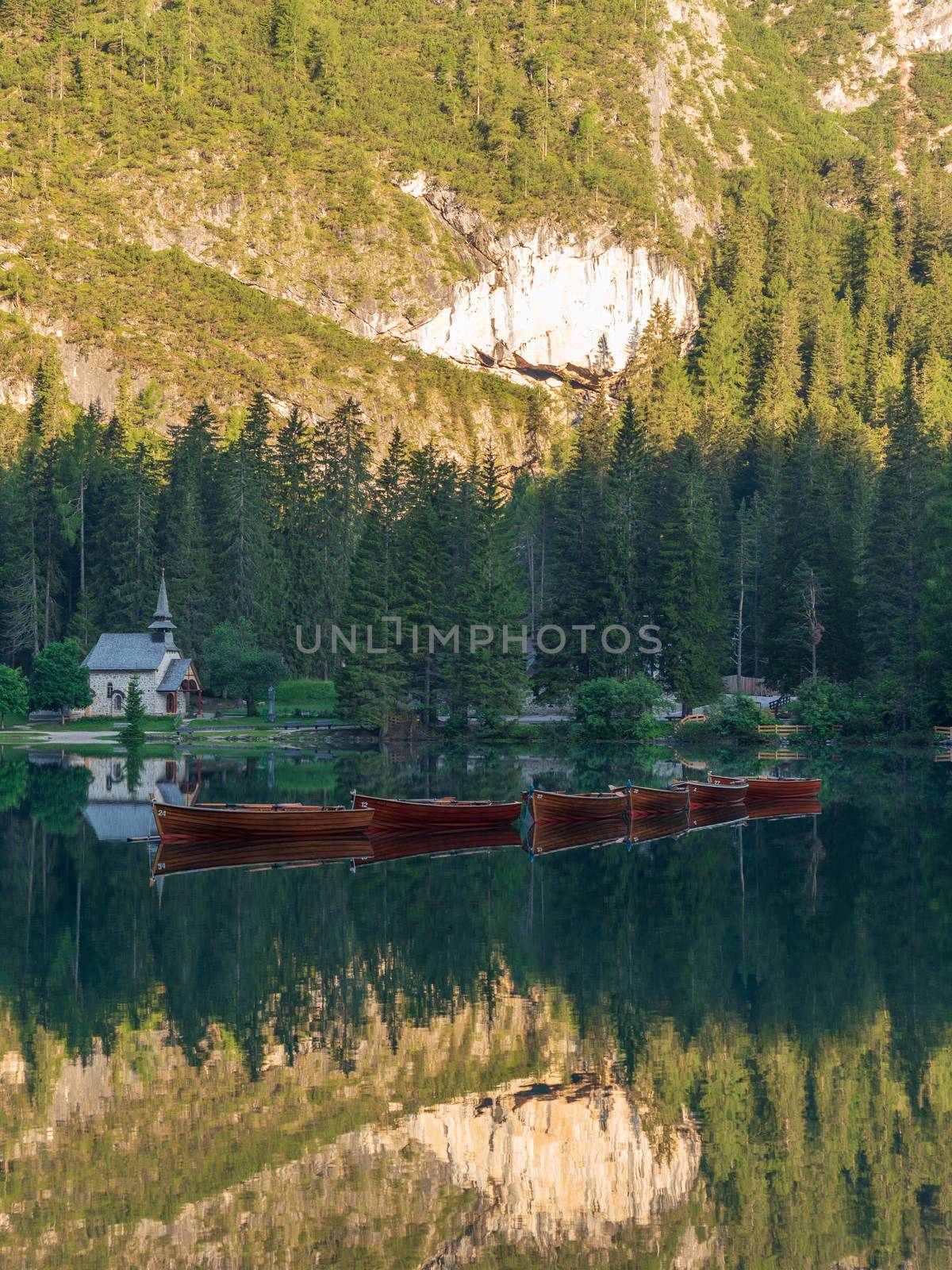 Row of wooden boats in front of a church reflected in the water of the mountain lake of Braies, landscape of the Italian dolomites