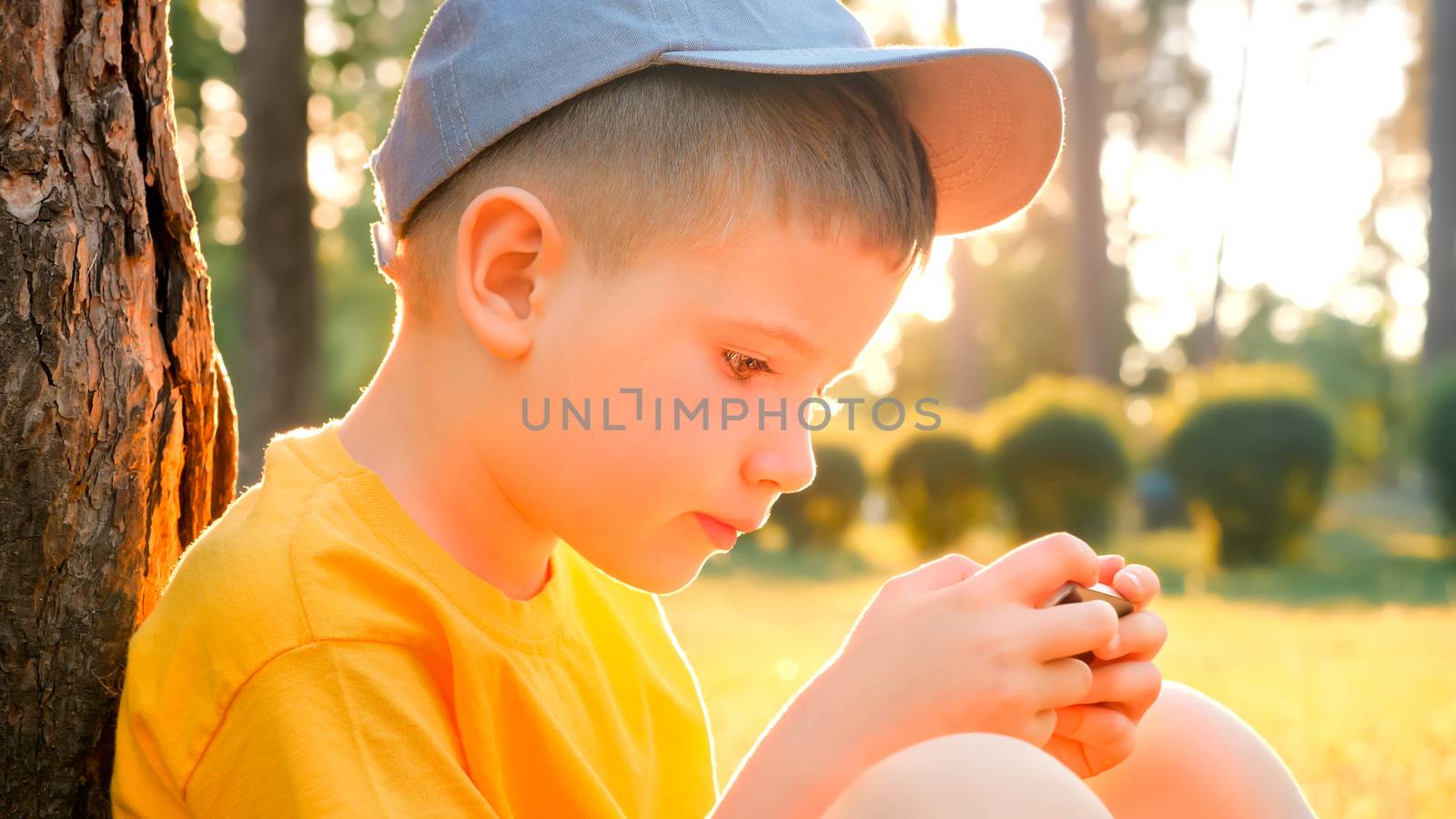 Child reading ebook on mobile outdoor. Kid with gadget in park, garden. Boy in cap playing game on smartphone. Elearning, education online. Preschooler spends time on fresh air. Pediatrics, psychology