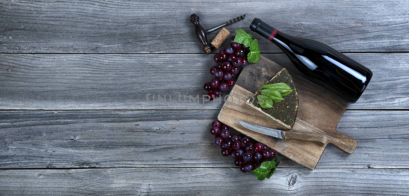 Fresh cheese wedge with a bottle of red wine plus basil leaves and grapes on rustic wood with copy space available 