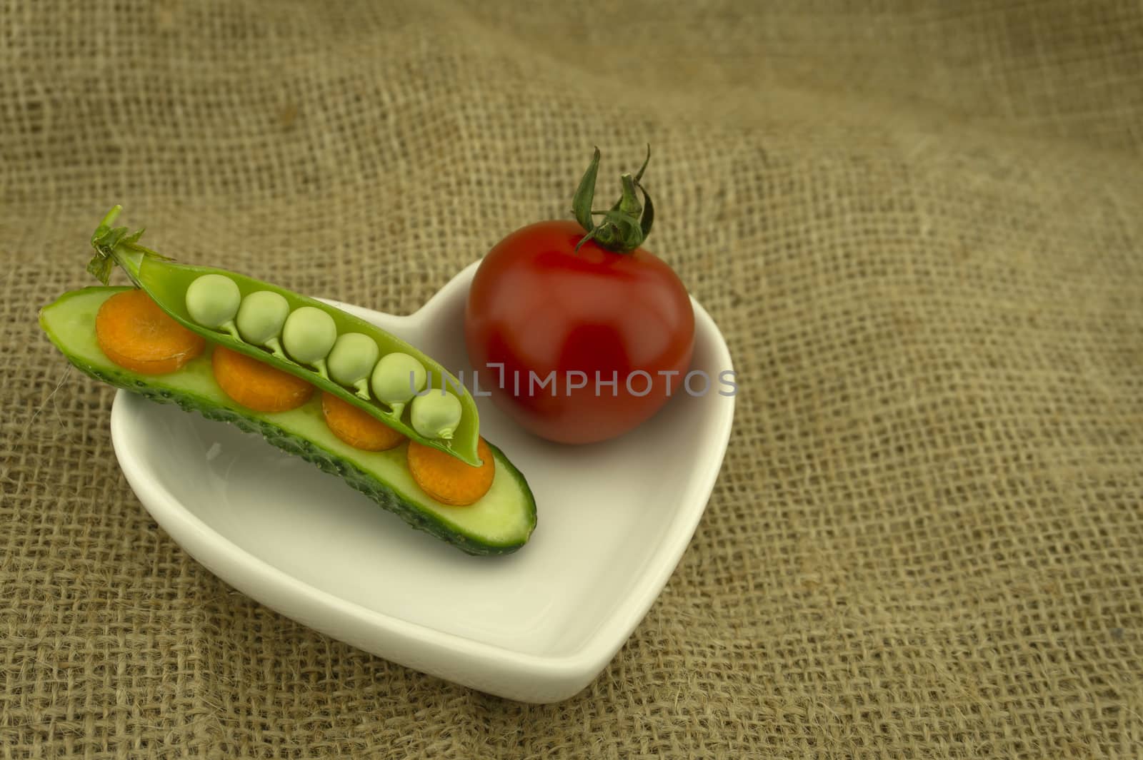 Healthy fresh vegetable sandwich with carrots, peas in a pod and cucumber balance on top of each and red ripe tomato in a heart-shaped plate on jute