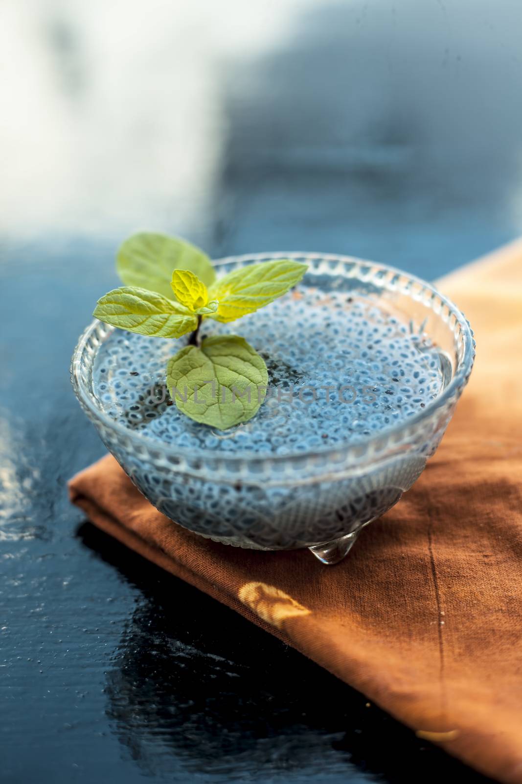 Close up of soaked sabja seeds or falooda seeds or sweet basil seeds in a glass bowl on brown colored napkin on wooden surface with some mint leaves in it.Used in many flavored beverages.