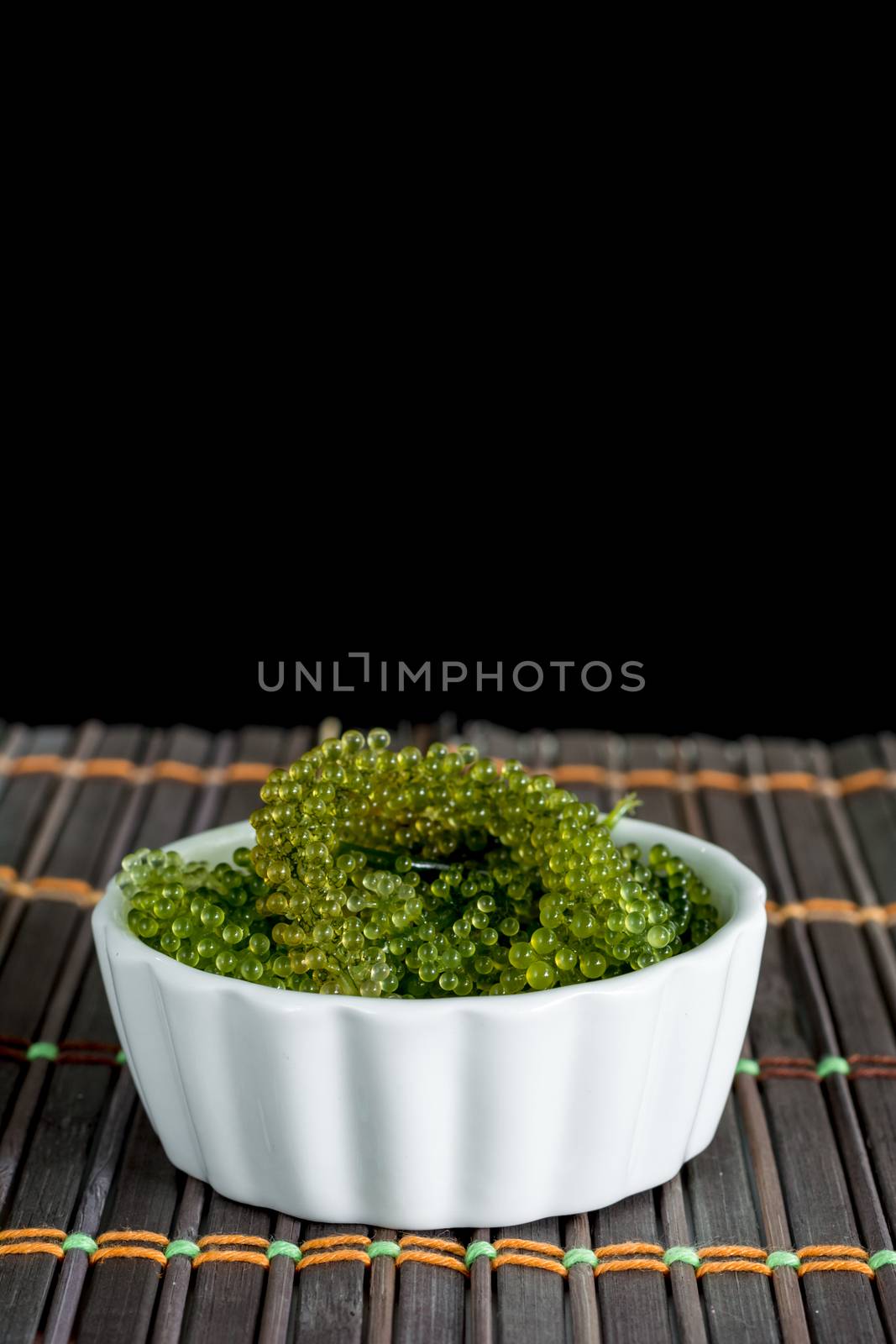 Fresh sea grapes or caviar seaweed in white bowl on wooden mat and black background. Healthy food concept.