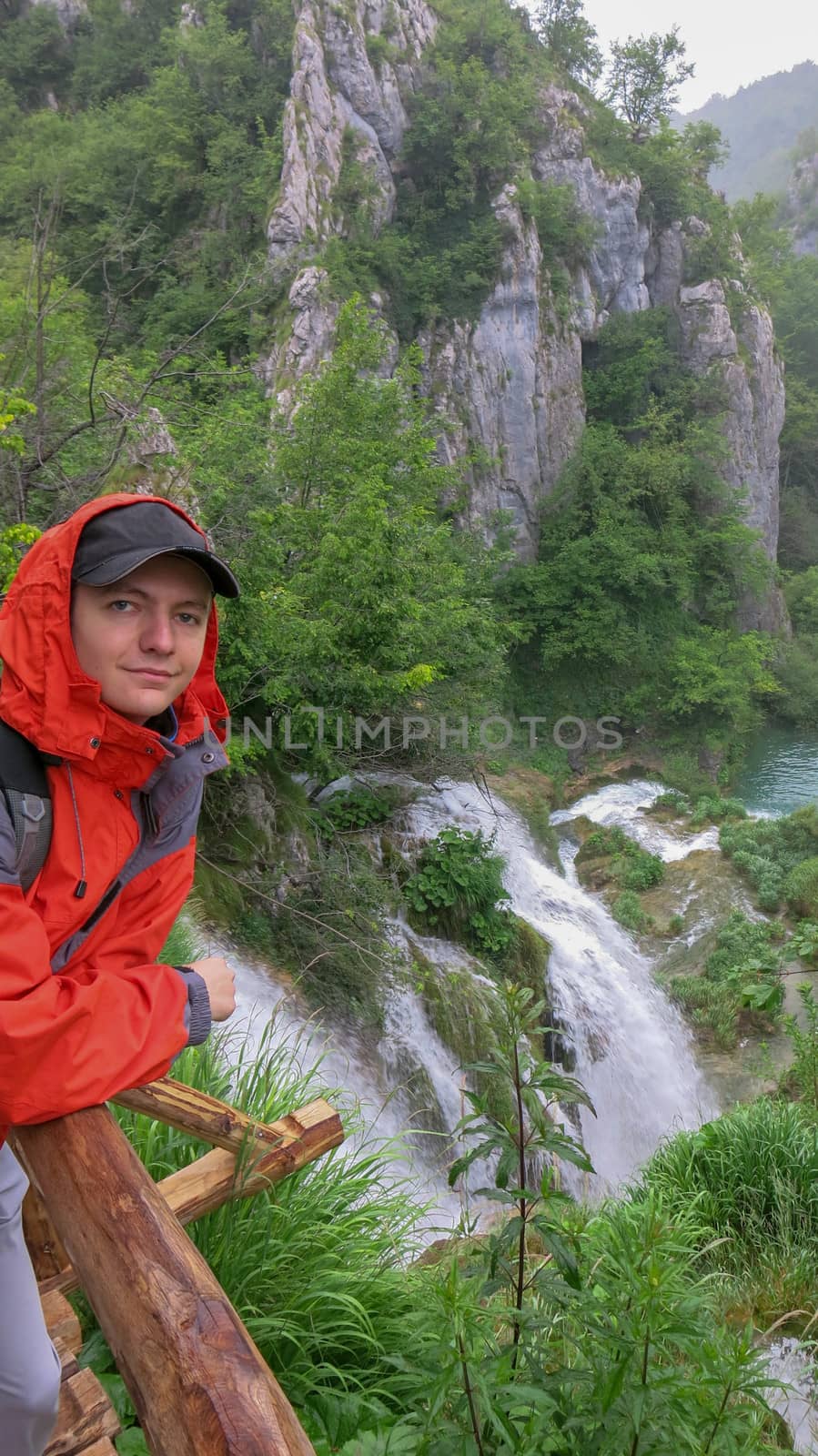 A young man leans on a wooden railing. In the background waterfalls in Plitvice Lakes National Park. Teenager in a red jacket stands against a waterfall.