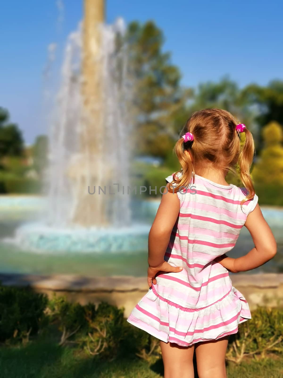 Preschooler little girl watching fountain rear view. Young female child enjoy being outside in summertime. Curiosity blonde looking at splashing droplets. enjoyment of holidays. Pretty kid photography