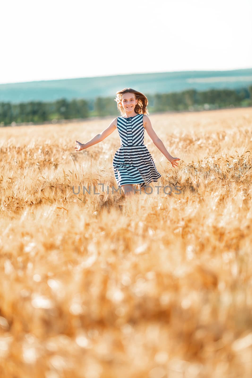 Happy teenage beautiful girl running down golden wheat field at the sunset