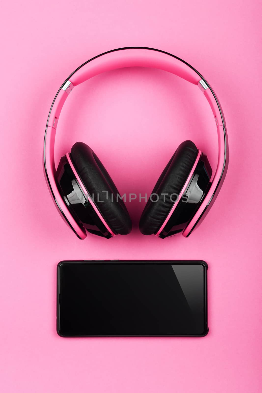 Close up modern wireless plastic pink headphones with big cushions and black mobile phone on table, elevated top view, directly above