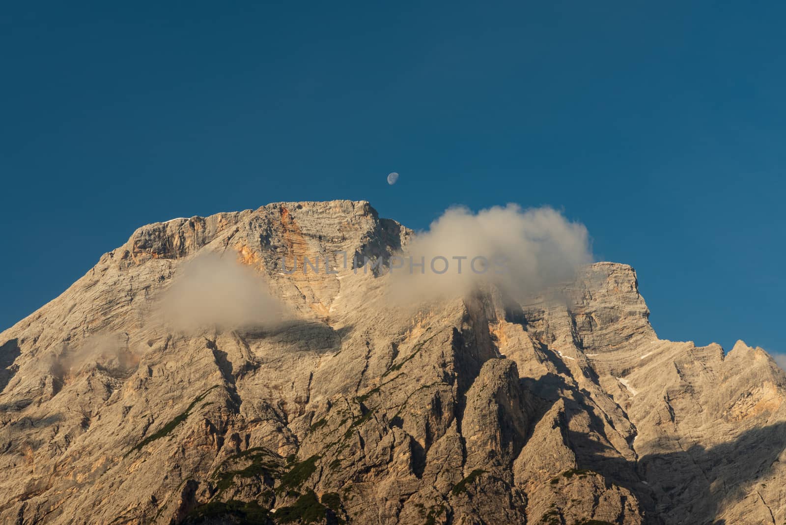 The top of the Seekofel with the shadow of two passing clouds and the small moon in the center of a blue sky, Italian Dolomites in South Tyrol