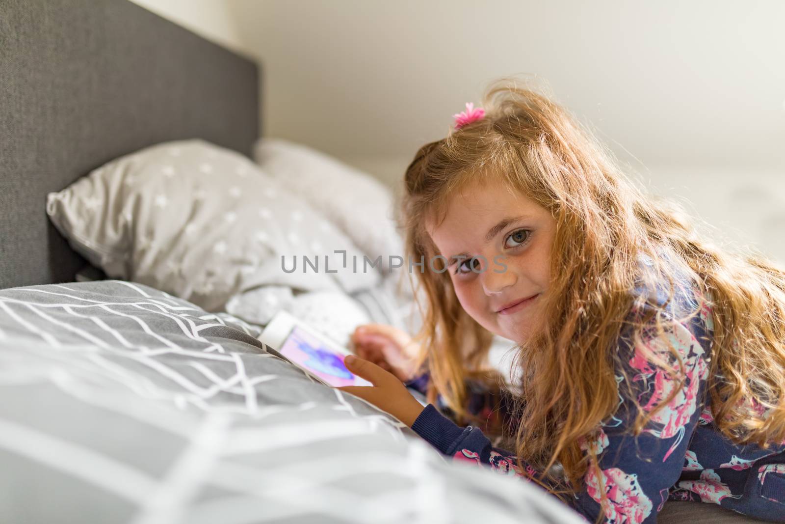 Smiling happy cute girl with long curly golden hair using digital tablet in bed.