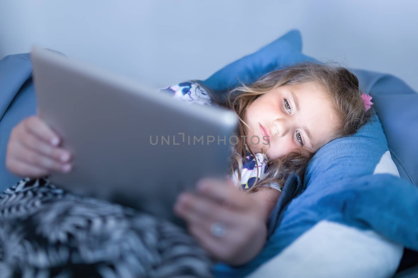 Focused small cute girls using digital tablet in bed, watching movie or play game in evening.