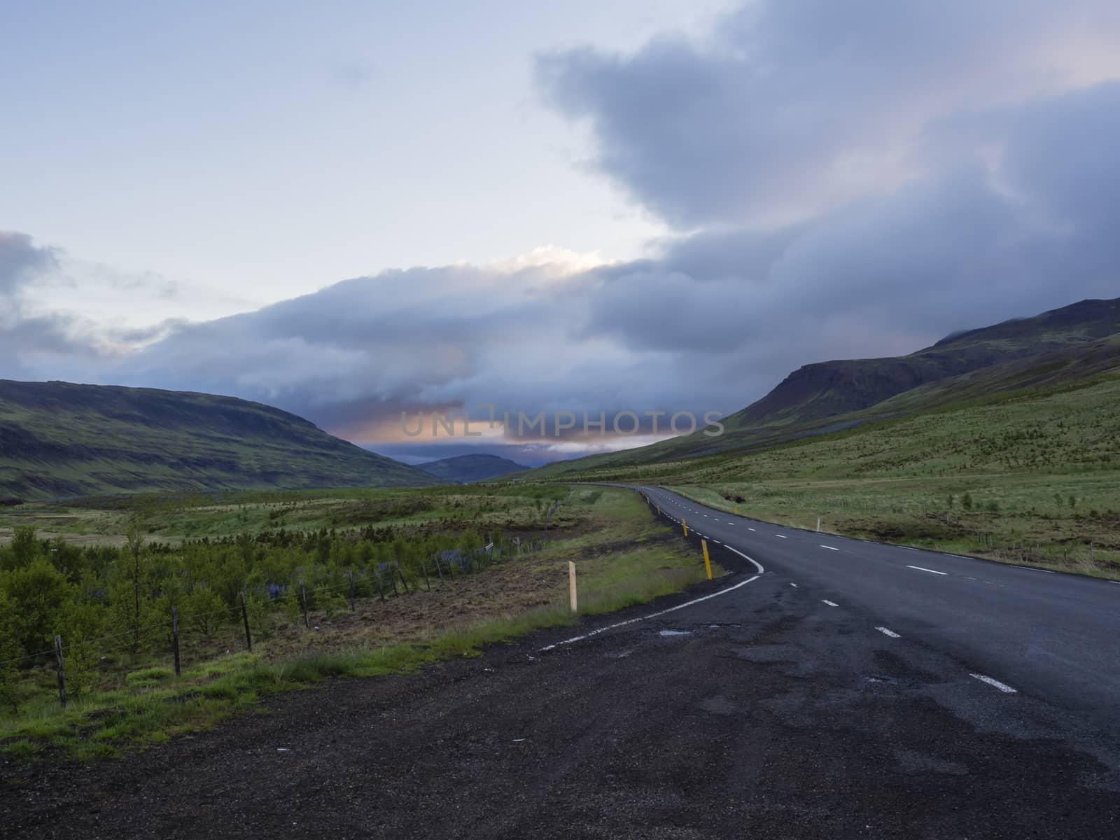 Asphalt road curve through empty northern landscape with green grass colorful hills and sunset dramatic sky, way to the mountains in Iceland western highlands, copy space